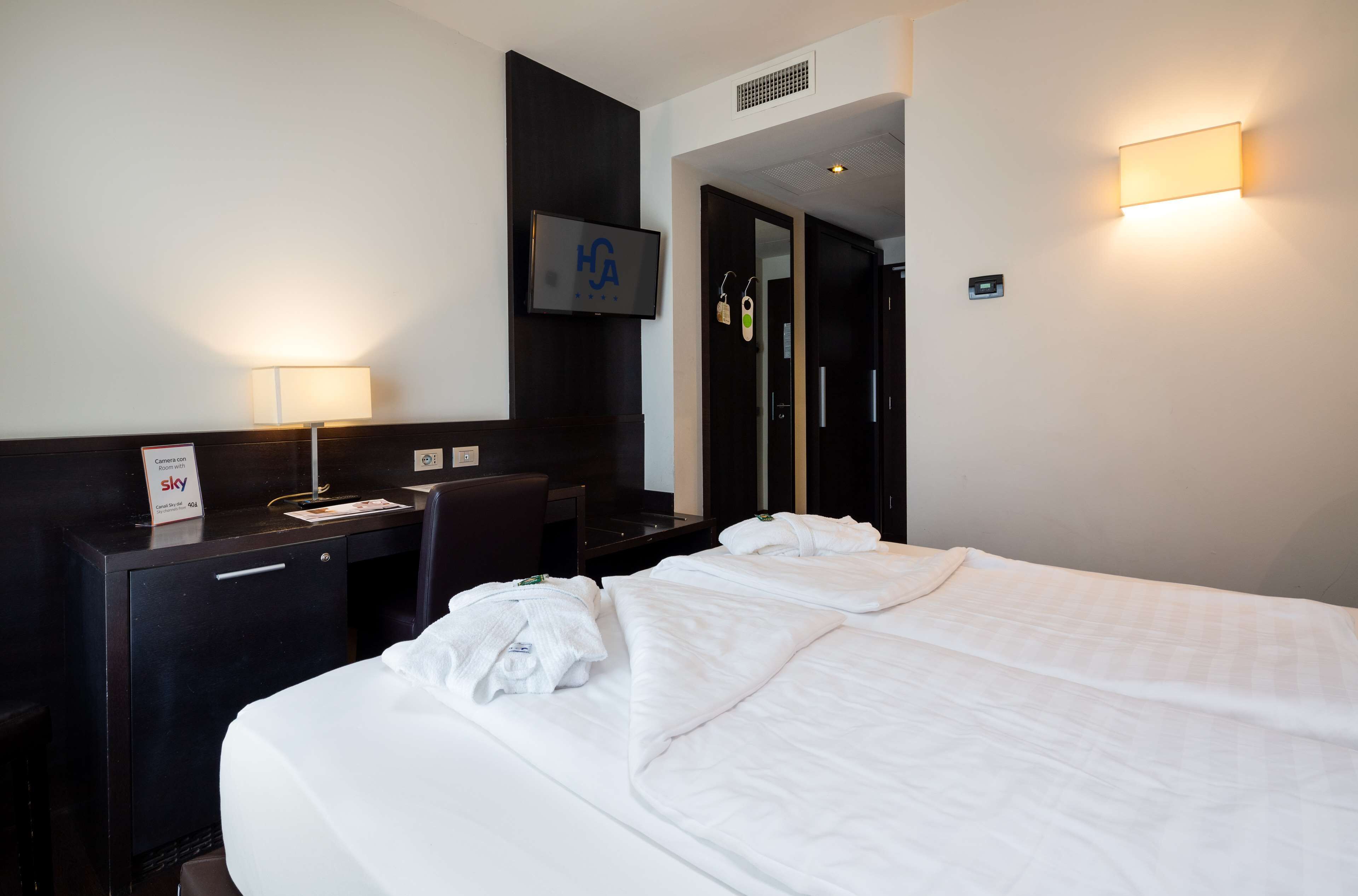A hotel room with a bed, a desk, a chair, a TV, and a wall-mounted light.