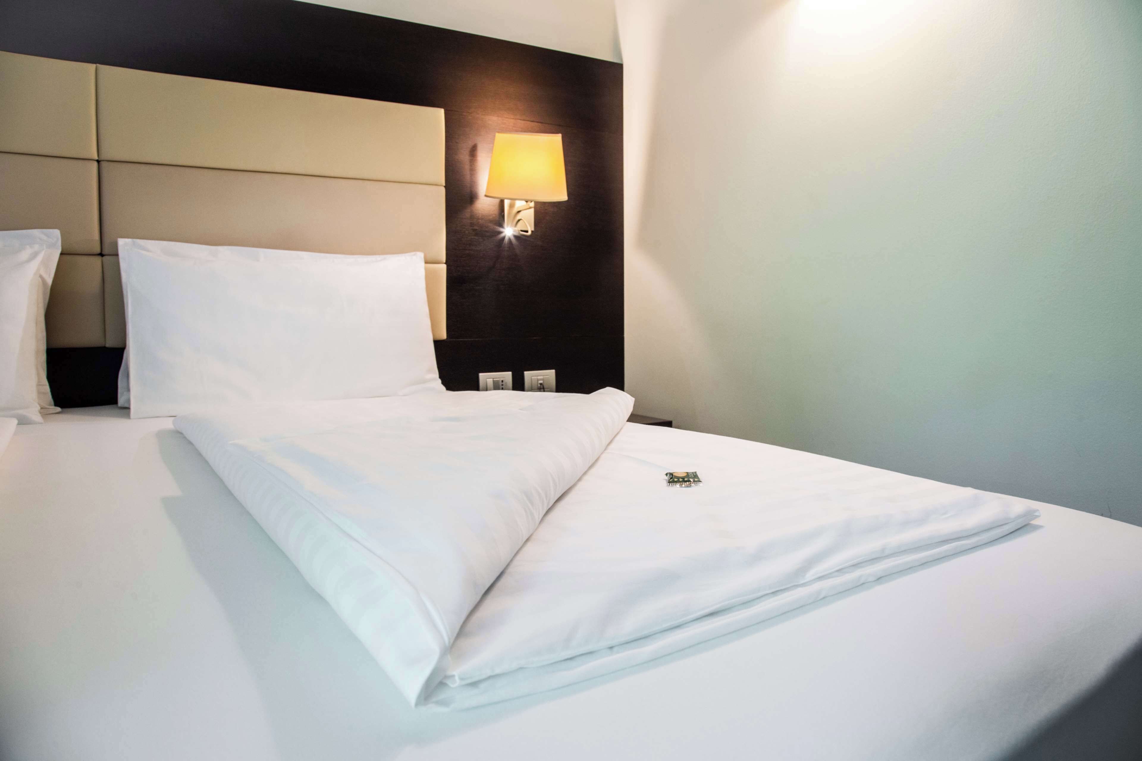 Una cama bien hecha con sábanas blancas, cabecera con lámpara y mesita de noche fijada a la pared con un aplique.