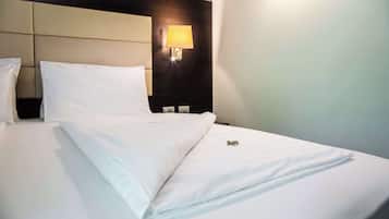 Una cama bien hecha con sábanas blancas, cabecera con lámpara y mesita de noche fijada a la pared con un aplique.