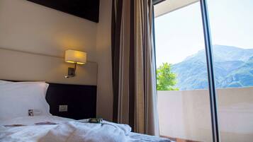 A hotel room with a bed, a nightstand, a wall-mounted lamp, and a large window offering a mountain view.