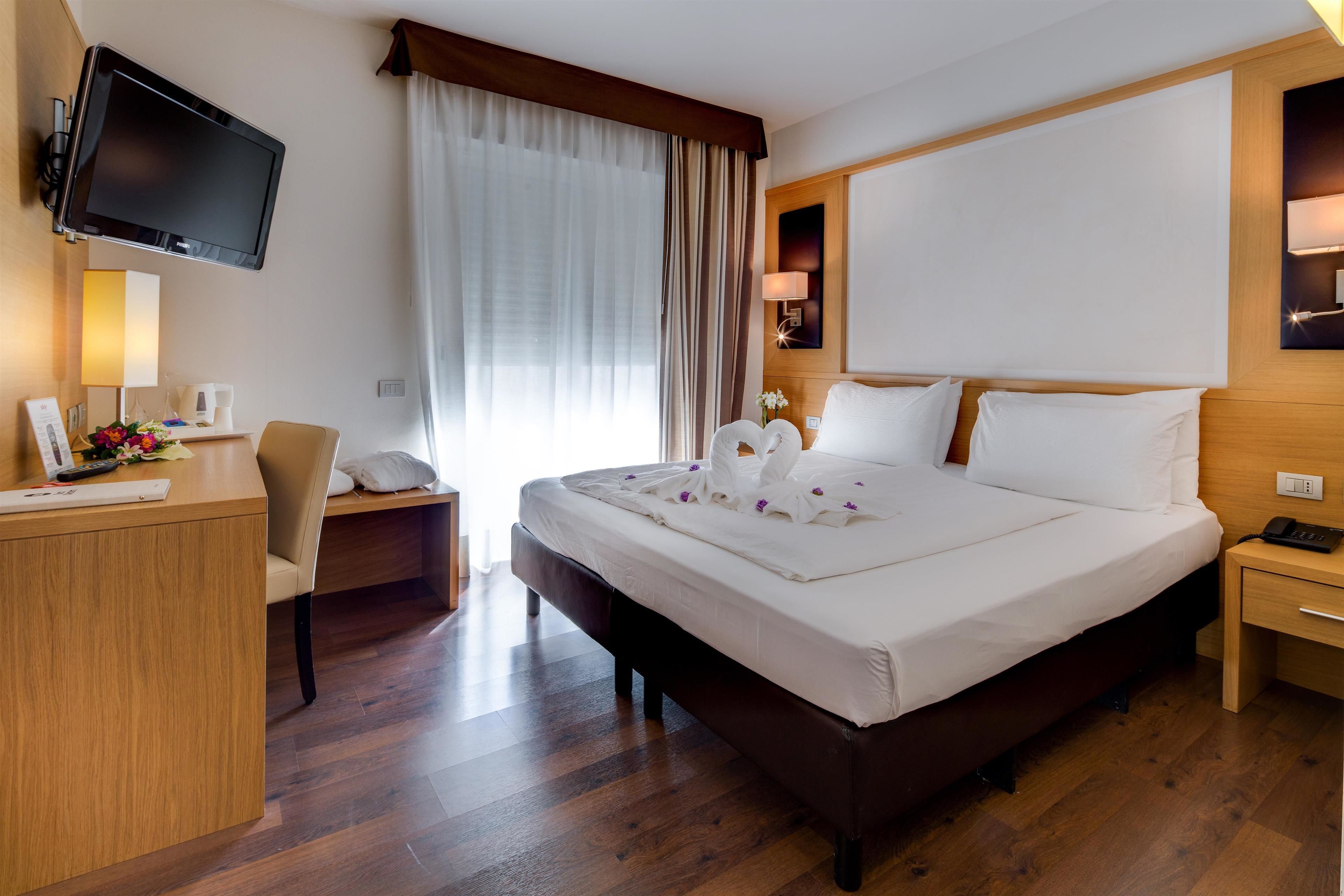 A hotel room with a large bed, a desk with a chair, a television mounted on the wall, and a bedside table with a lamp.