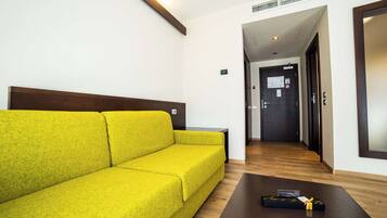 A hotel room with a bright yellow sofa, a wooden coffee table, and a dark door at the end of a hallway.