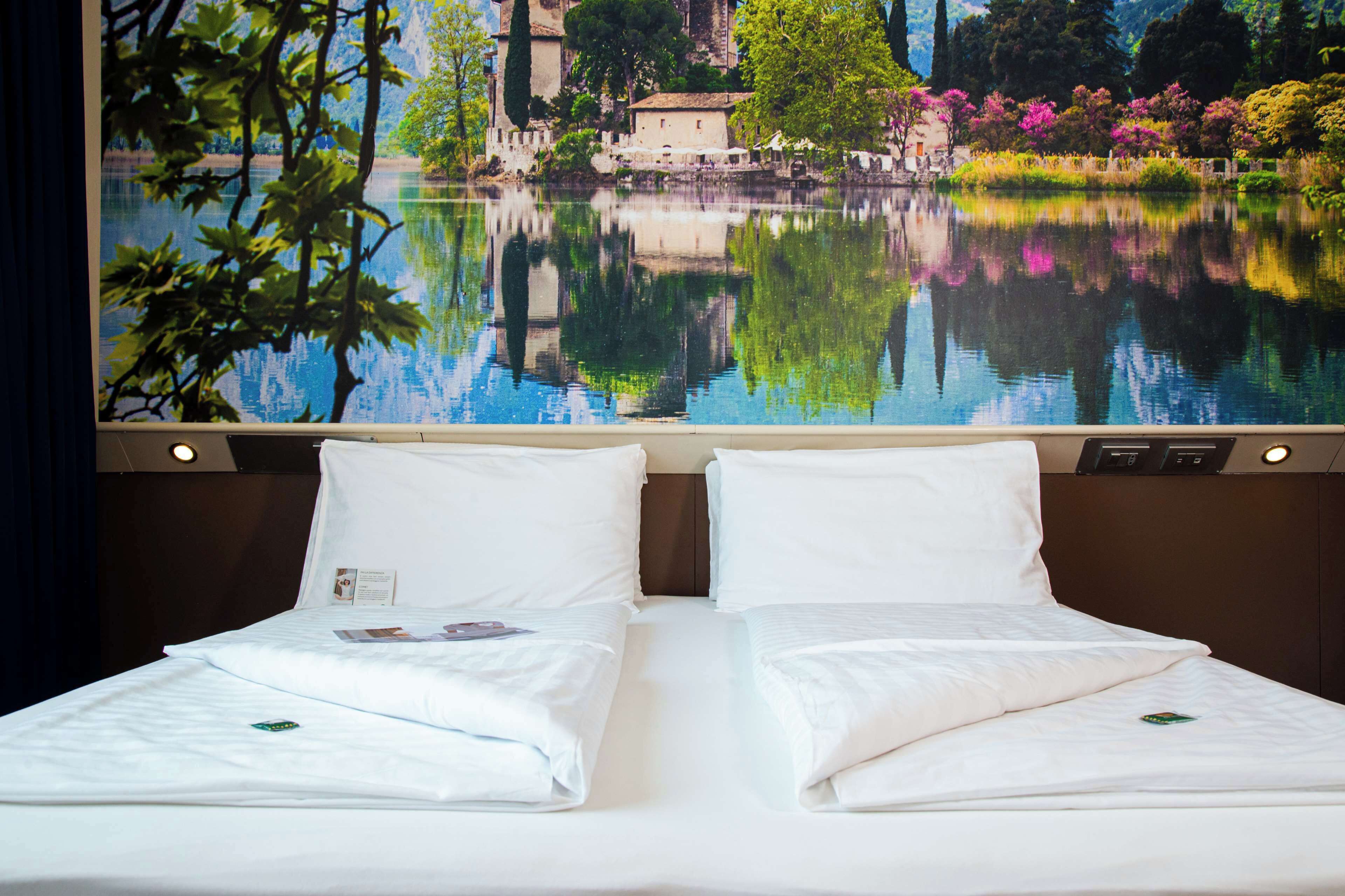 Habitación de hotel con dos camas, un ventanal con vistas a un lago y edificios, y una mesita de noche con lámpara.