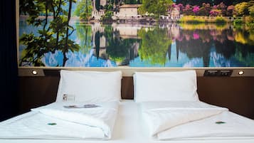 Habitación de hotel con dos camas, un ventanal con vistas a un lago y edificios, y una mesita de noche con lámpara.
