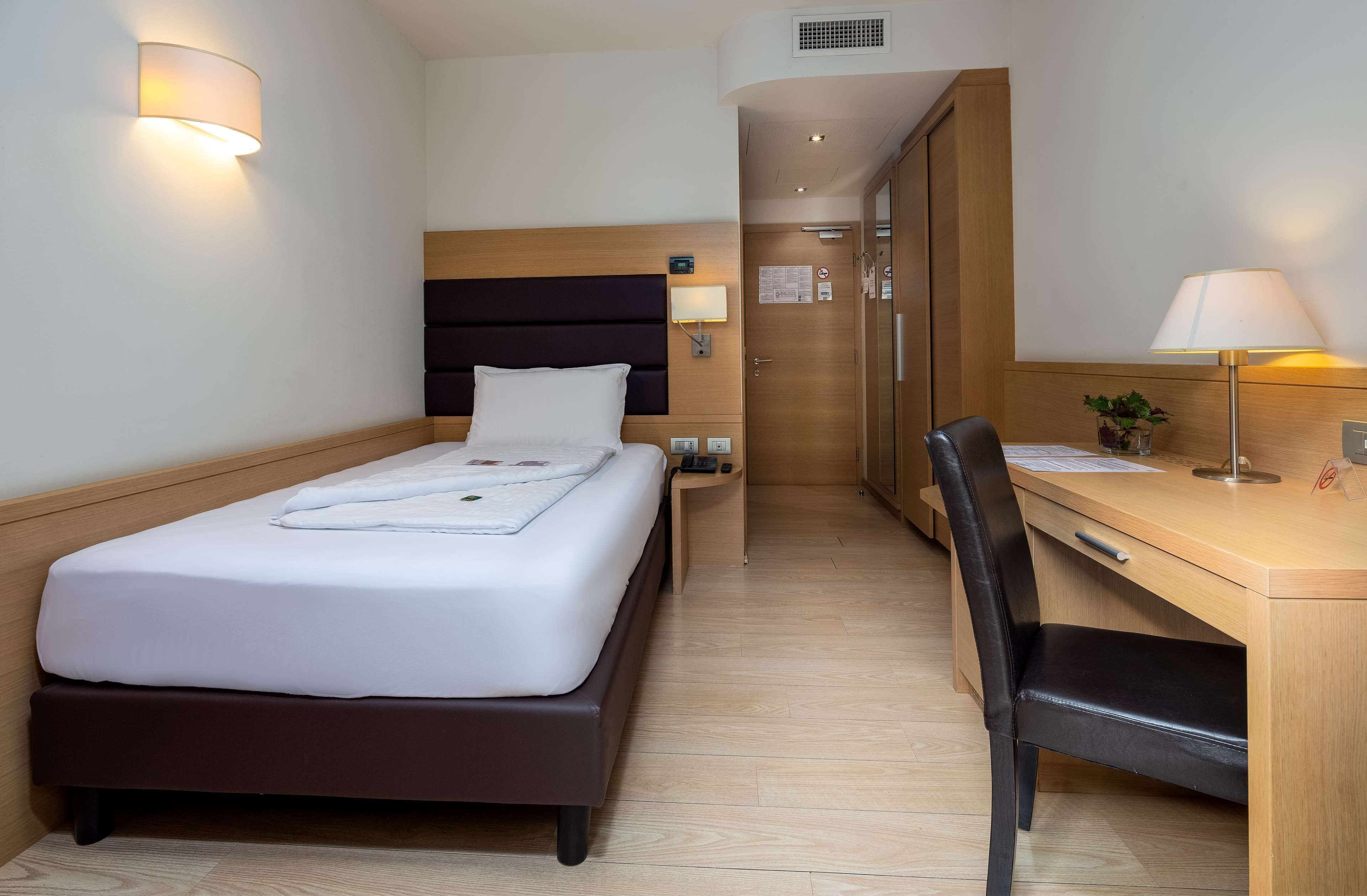 A hotel room with a bed, bedside table, lamp, desk, chair, and a small potted plant.