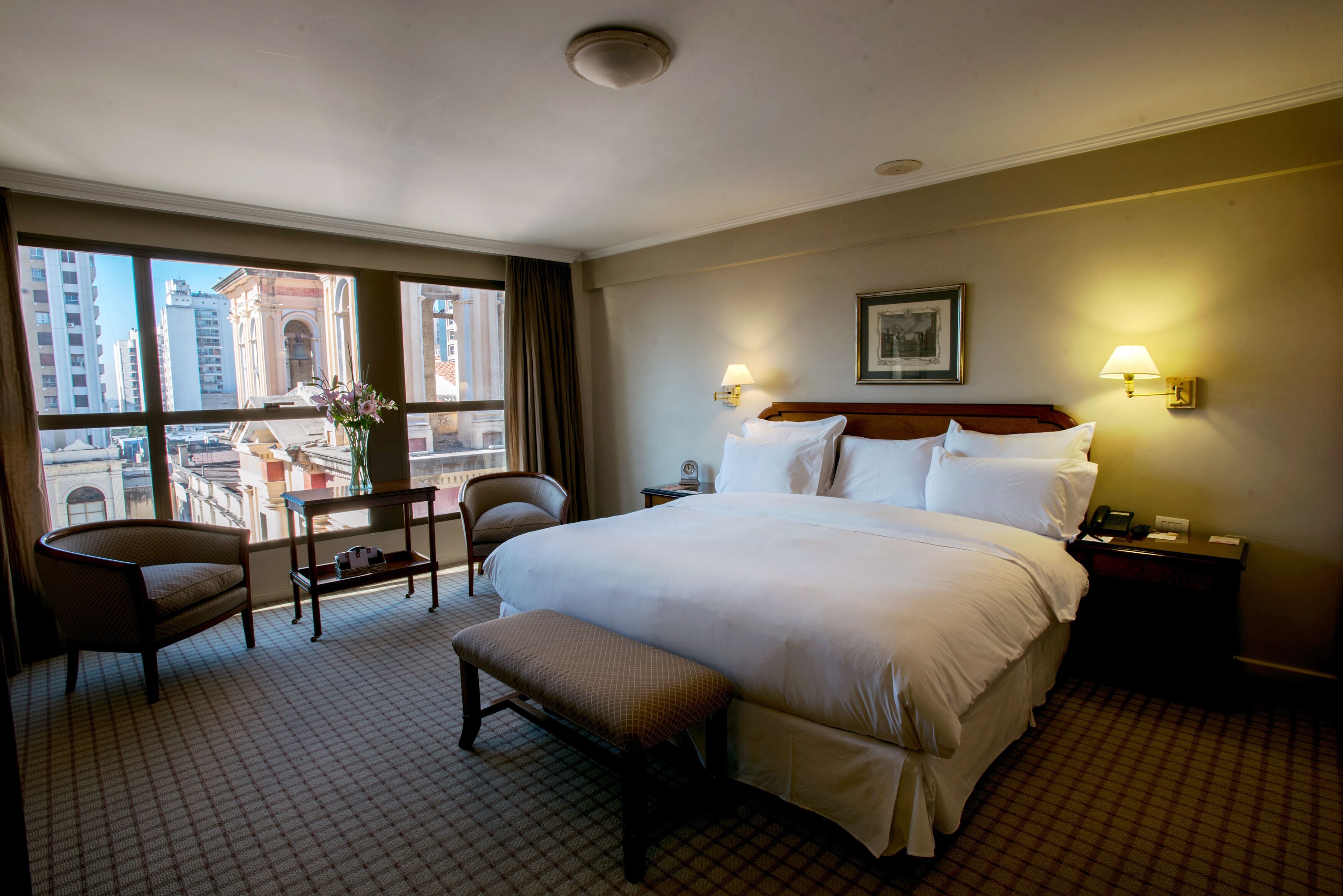 A hotel room with a large bed, a chair, a small table, and a bench. There is a window with a view of buildings outside.