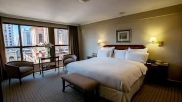 A hotel room with a large bed, a chair, a small table, and a bench. There is a window with a view of buildings outside.