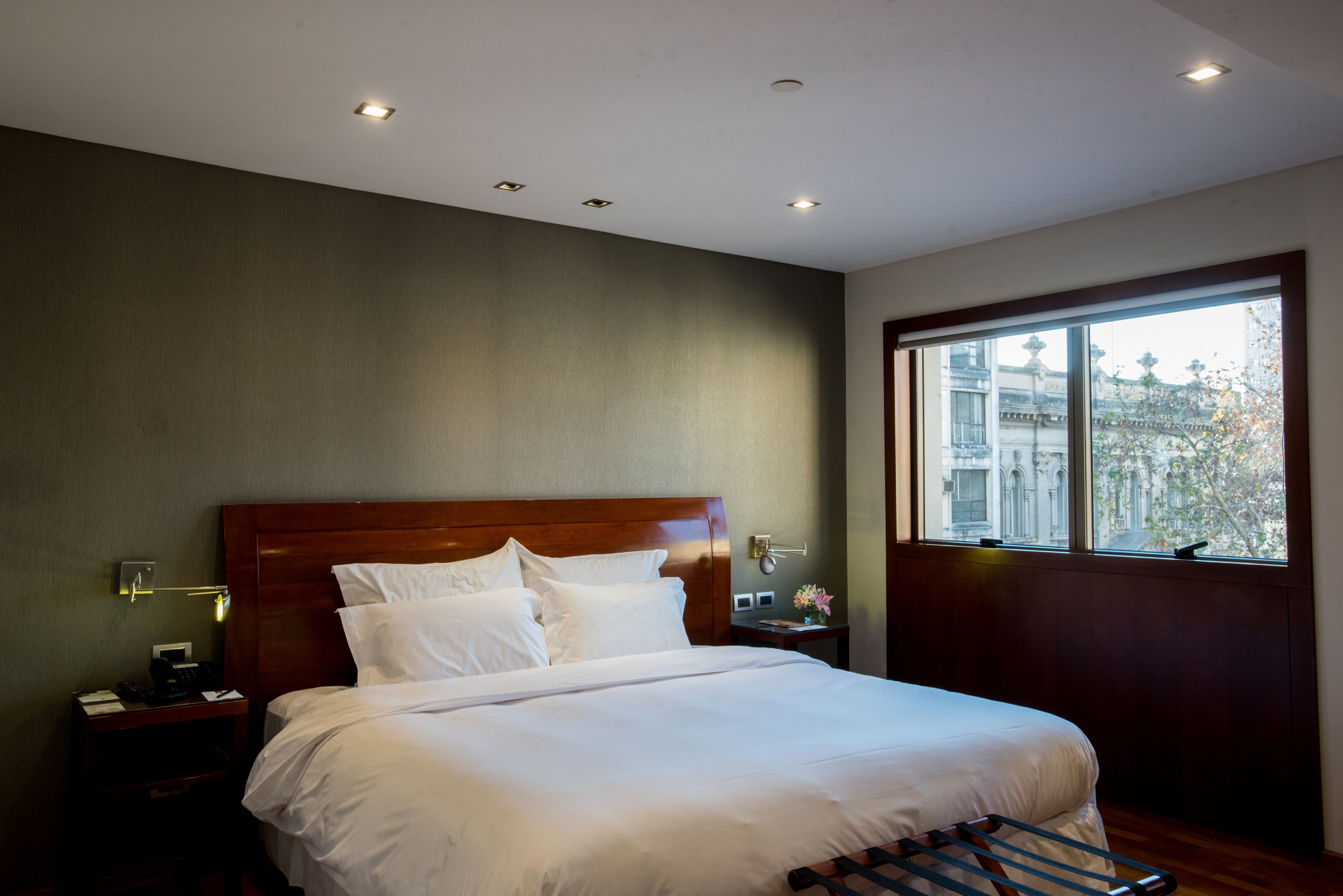 Ein Hotelzimmer mit einem großen Bett, einem Holz-Kopfstück, einem Nachttisch mit Lampe und einem Fenster mit Blick auf Gebäude.
