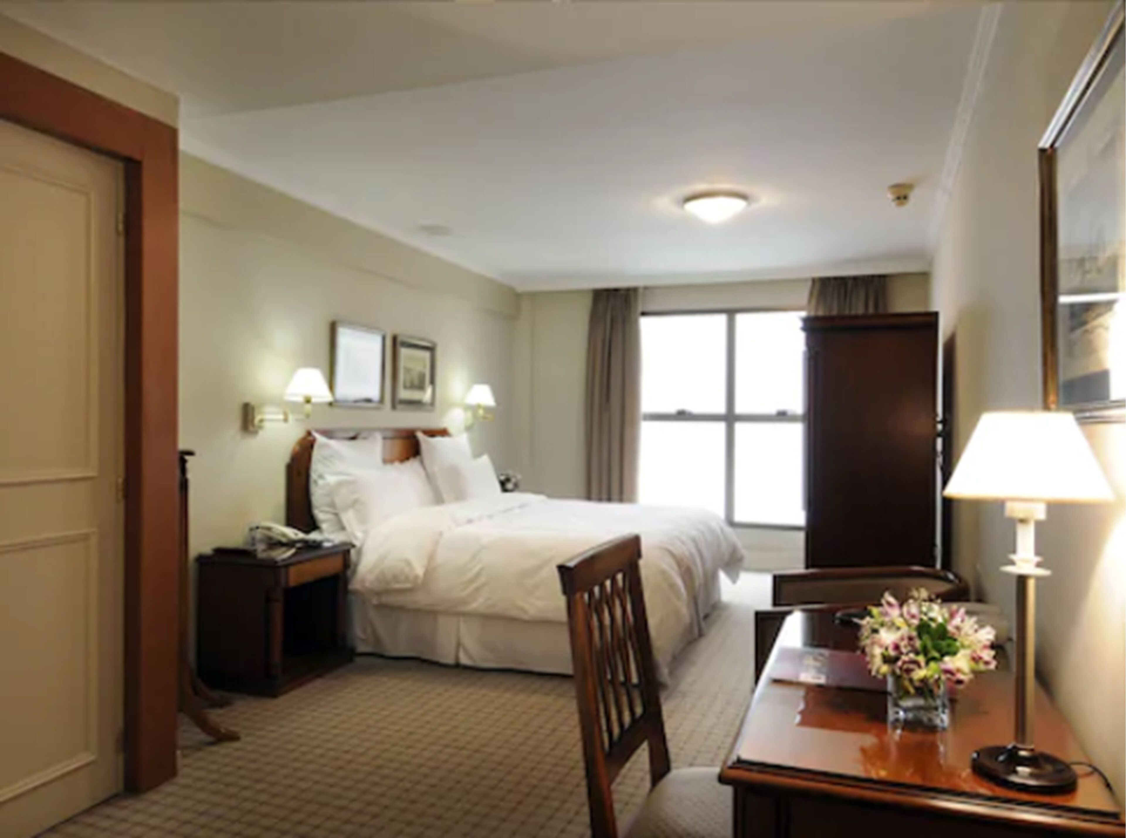 Una habitación de hotel con cama, mesita de noche, lámpara, silla y ventana con cortinas.