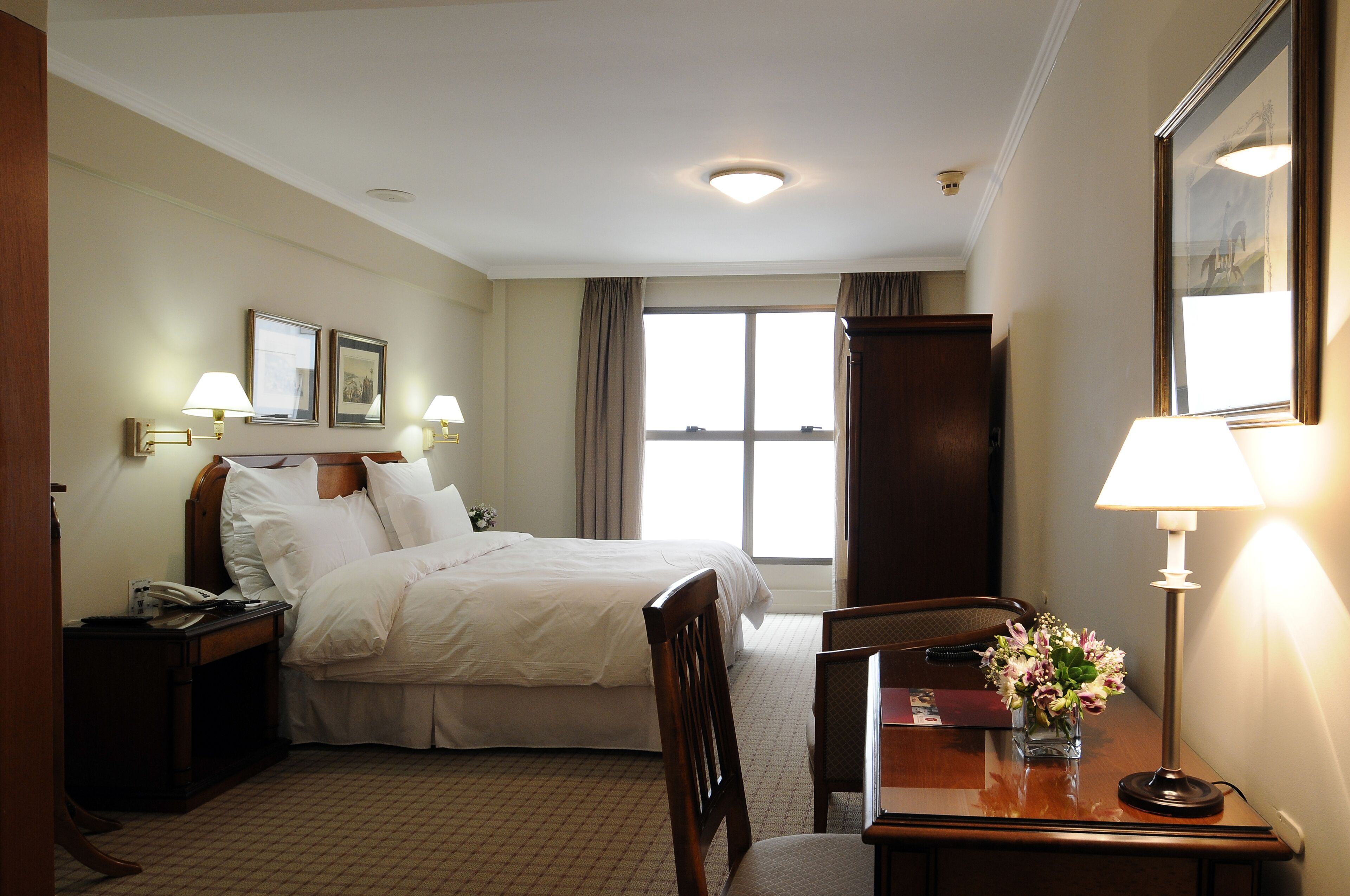 A hotel room with a neatly made bed, a desk with a lamp, a chair, a nightstand with a phone, and a window with curtains.