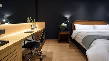 Habitación de hotel con un escritorio de madera, una silla de oficina negra, una cama con ropa de cama blanca y una mesita de noche con lámpara.