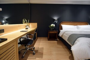 Habitación de hotel con un escritorio de madera, una silla de oficina negra, una cama con ropa de cama blanca y una mesita de noche con lámpara.