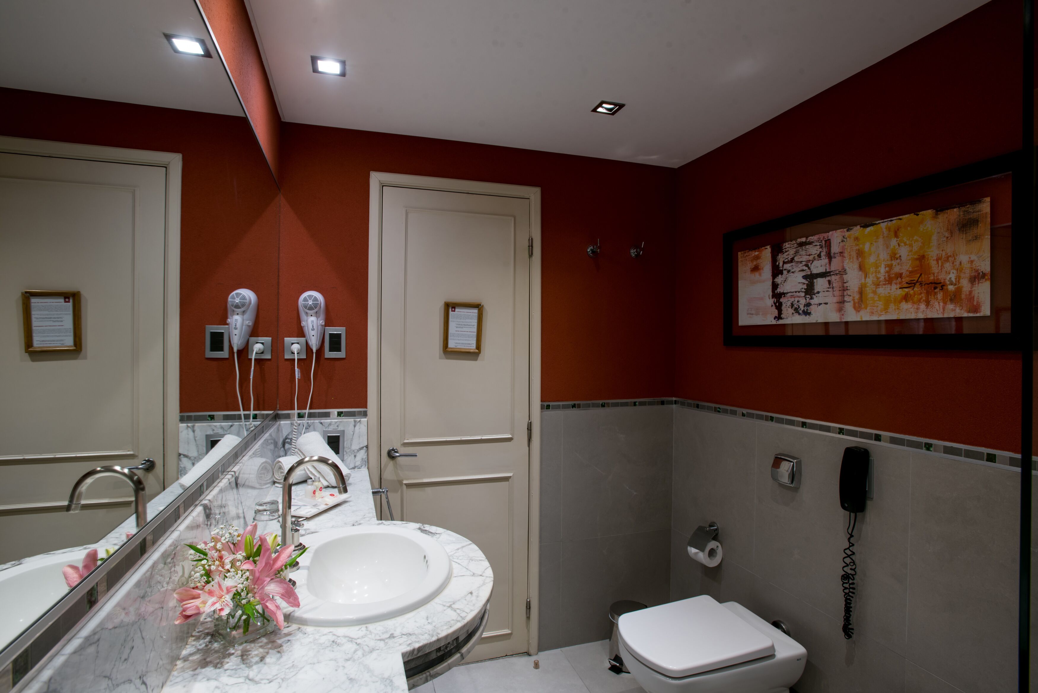 Un baño con un espejo grande, dos secadores de pelo, un lavabo con encimera de mármol, un inodoro y un teléfono en la pared.
