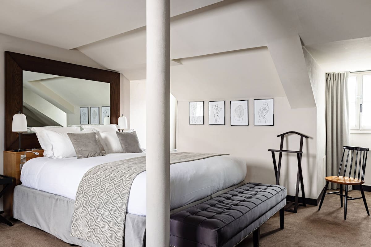 Une chambre à coucher avec un grand lit, un banc, une chaise et un miroir. Des tableaux encadrés ornent le mur.