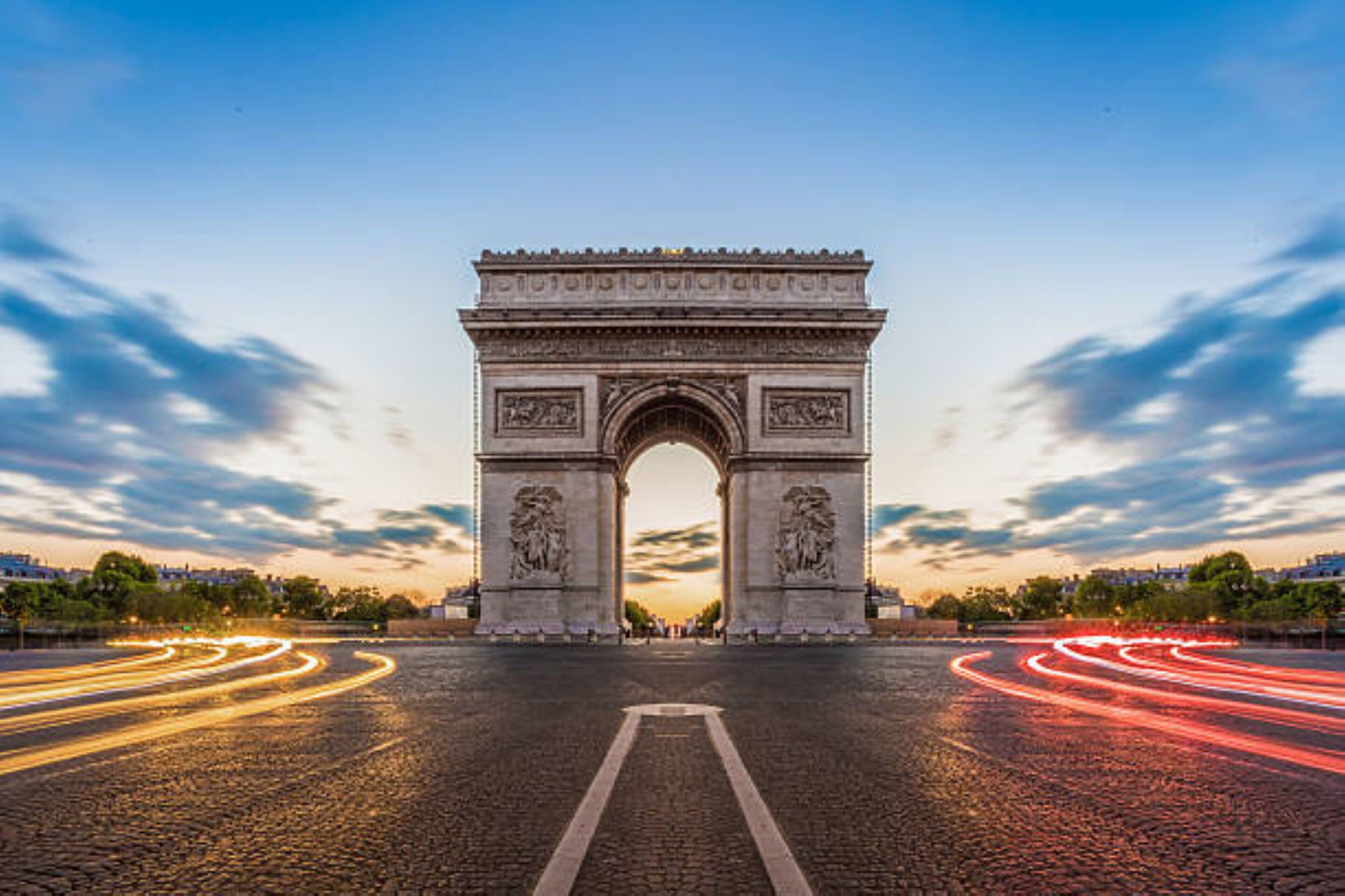 A large stone archway with intricate carvings, leading to a road with light trails indicating traffic.