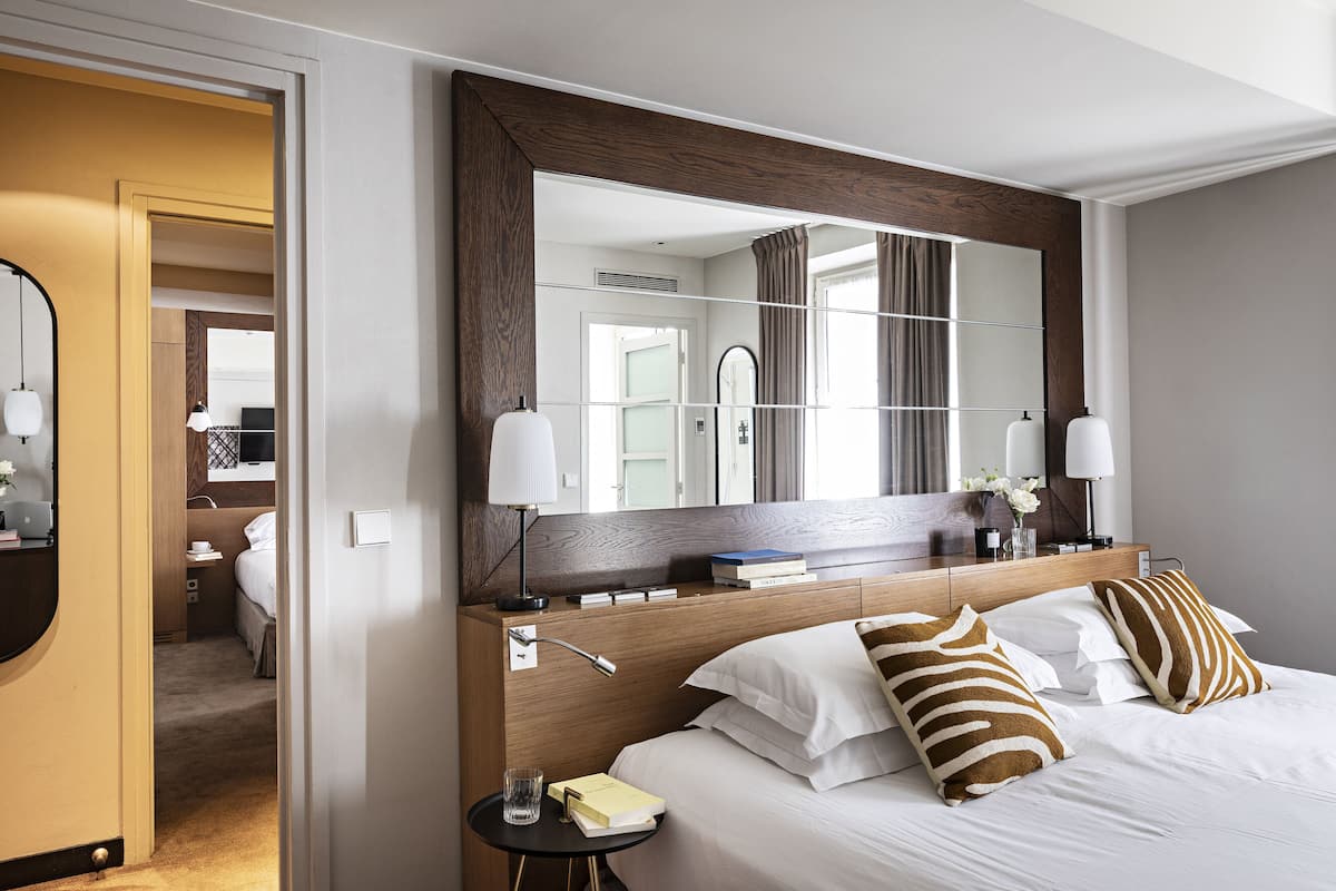 Une chambre d’hôtel avec un grand miroir, une tête de lit en bois, du linge de lit blanc et une table de chevet avec une lampe et des livres.