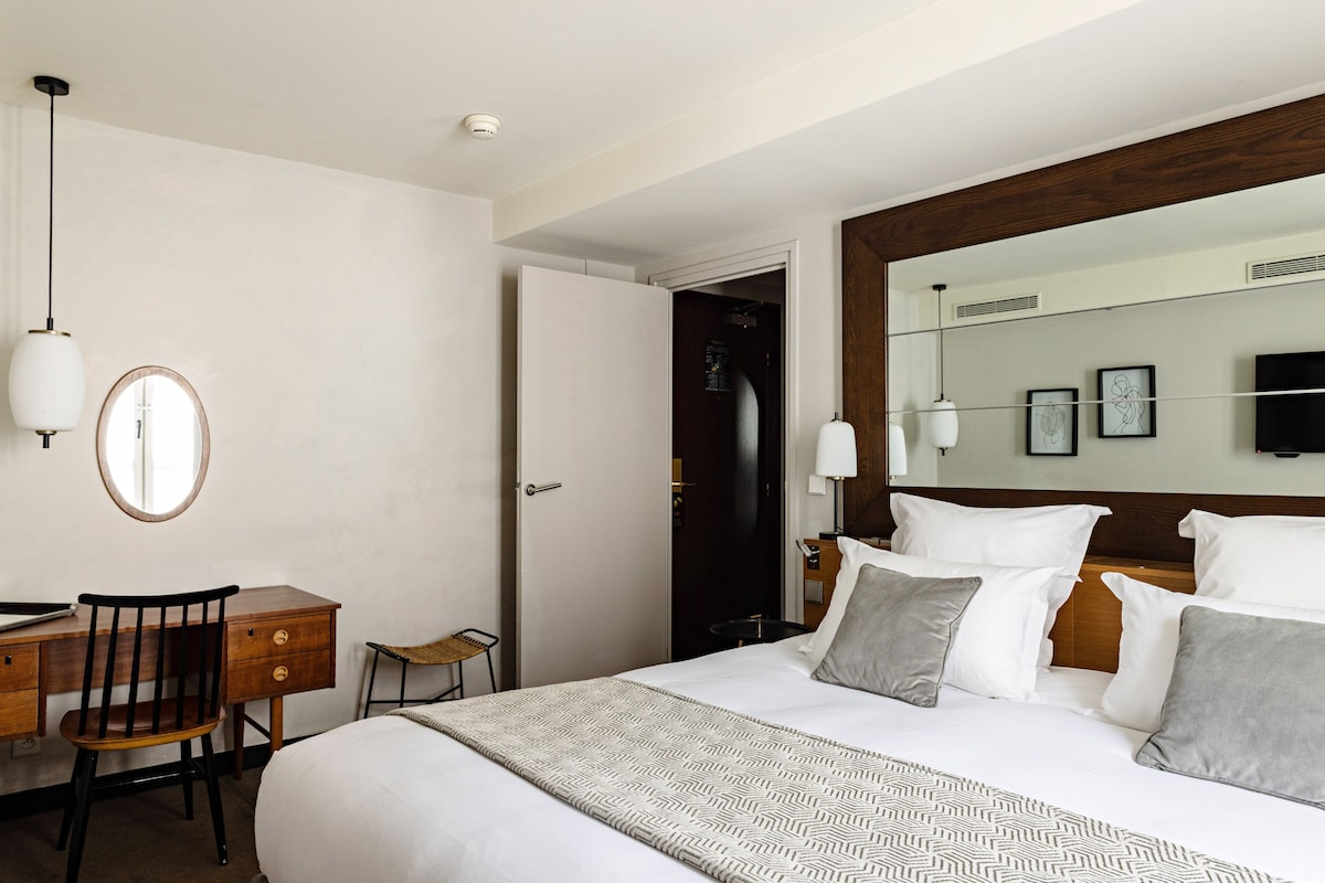 A hotel room with a bed, a desk, a chair, a mirror, and a door leading to another room.