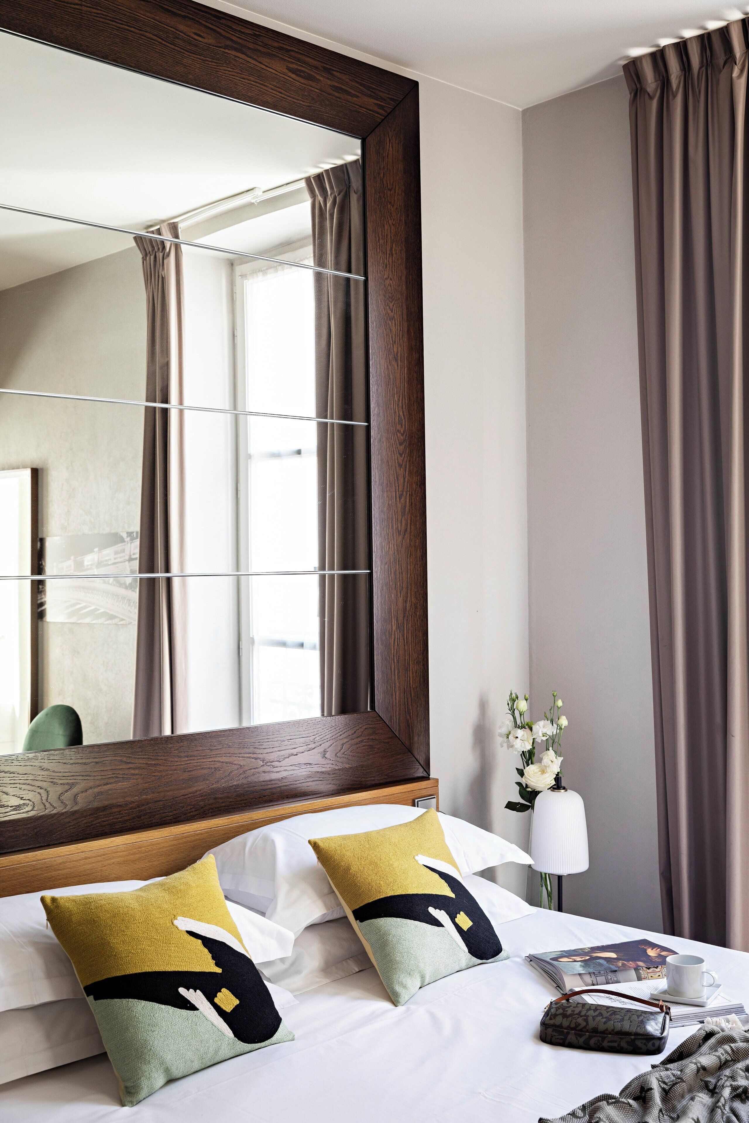 A hotel room with a large mirror, a bed with multiple pillows, a bedside table with a lamp, and a window with curtains.