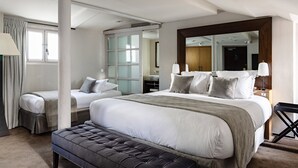 Habitación de hotel moderna con una cama grande, otra más pequeña, un banco, un espejo y un baño visible a través de una puerta de cristal.