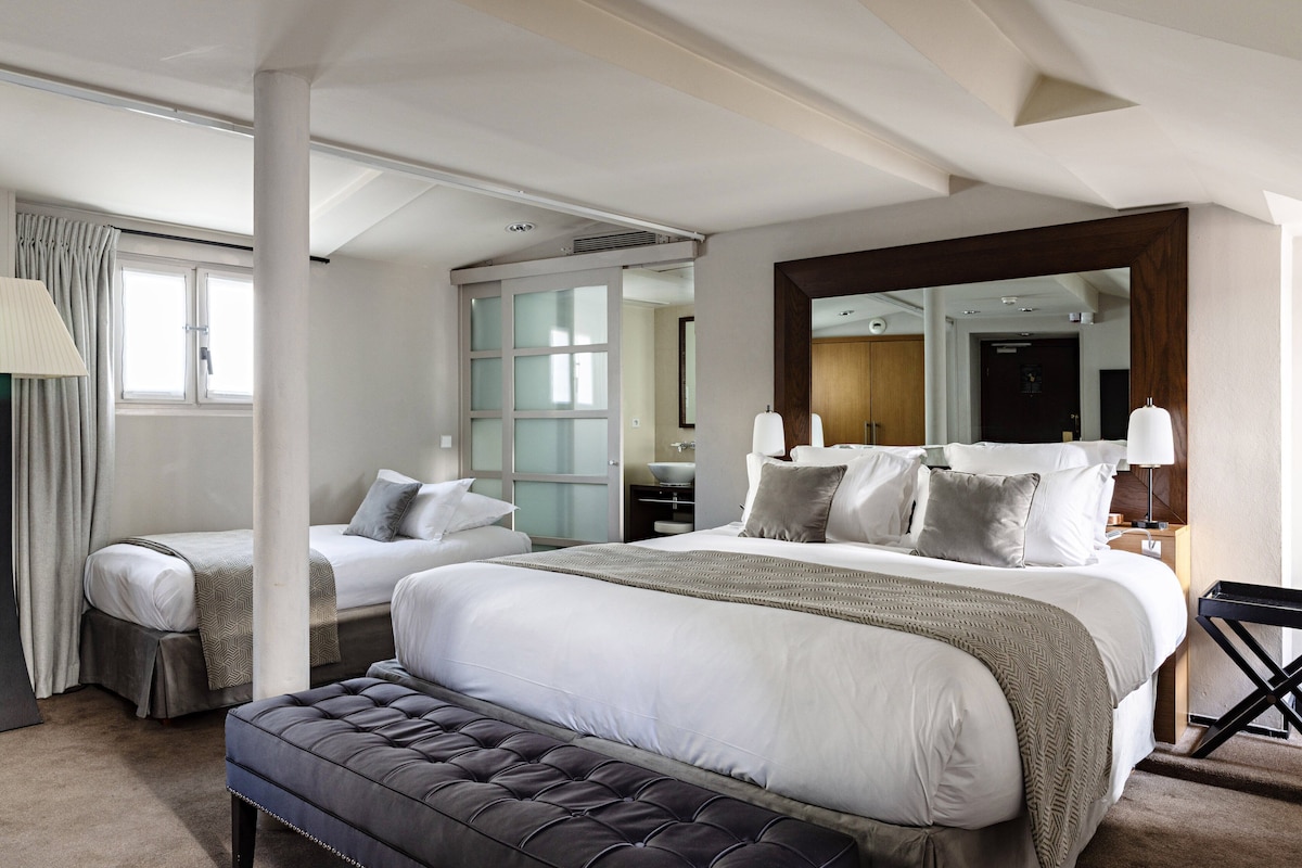 Habitación de hotel moderna con una cama grande, otra más pequeña, un banco, un espejo y un baño visible a través de una puerta de cristal.