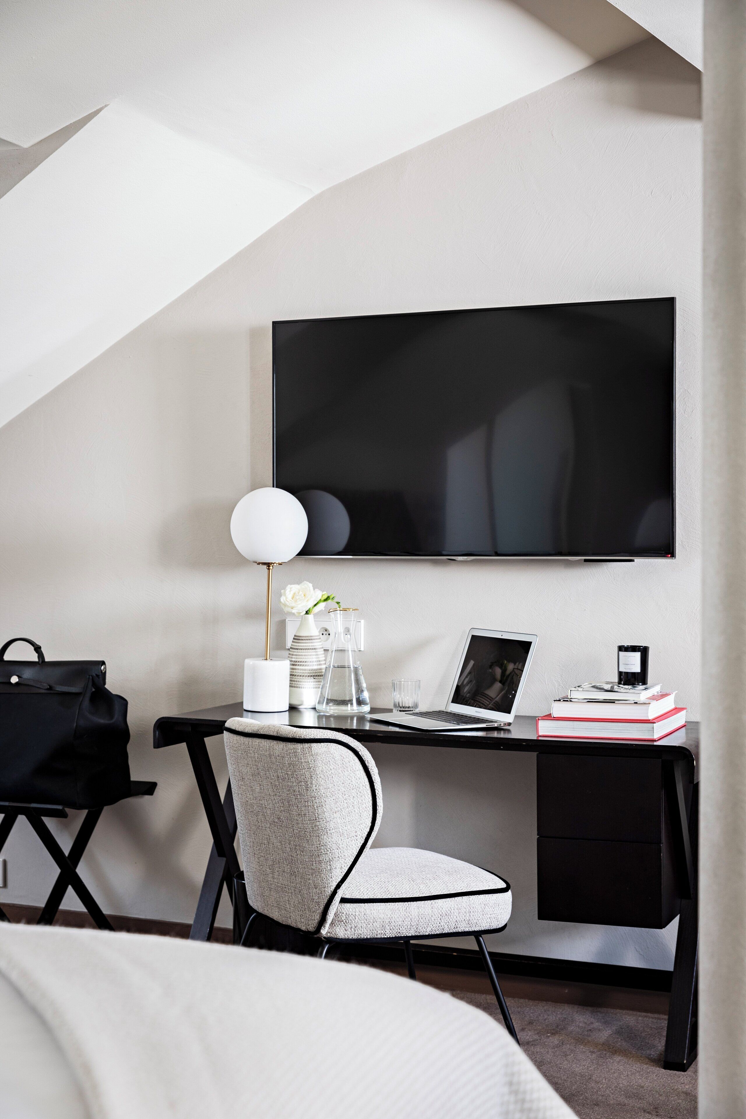 Habitación de hotel moderna con un escritorio, una silla, un televisor de pantalla plana en la pared, una computadora portátil, un jarrón con flores, una lámpara, una bolsa negra y una pila de libros.