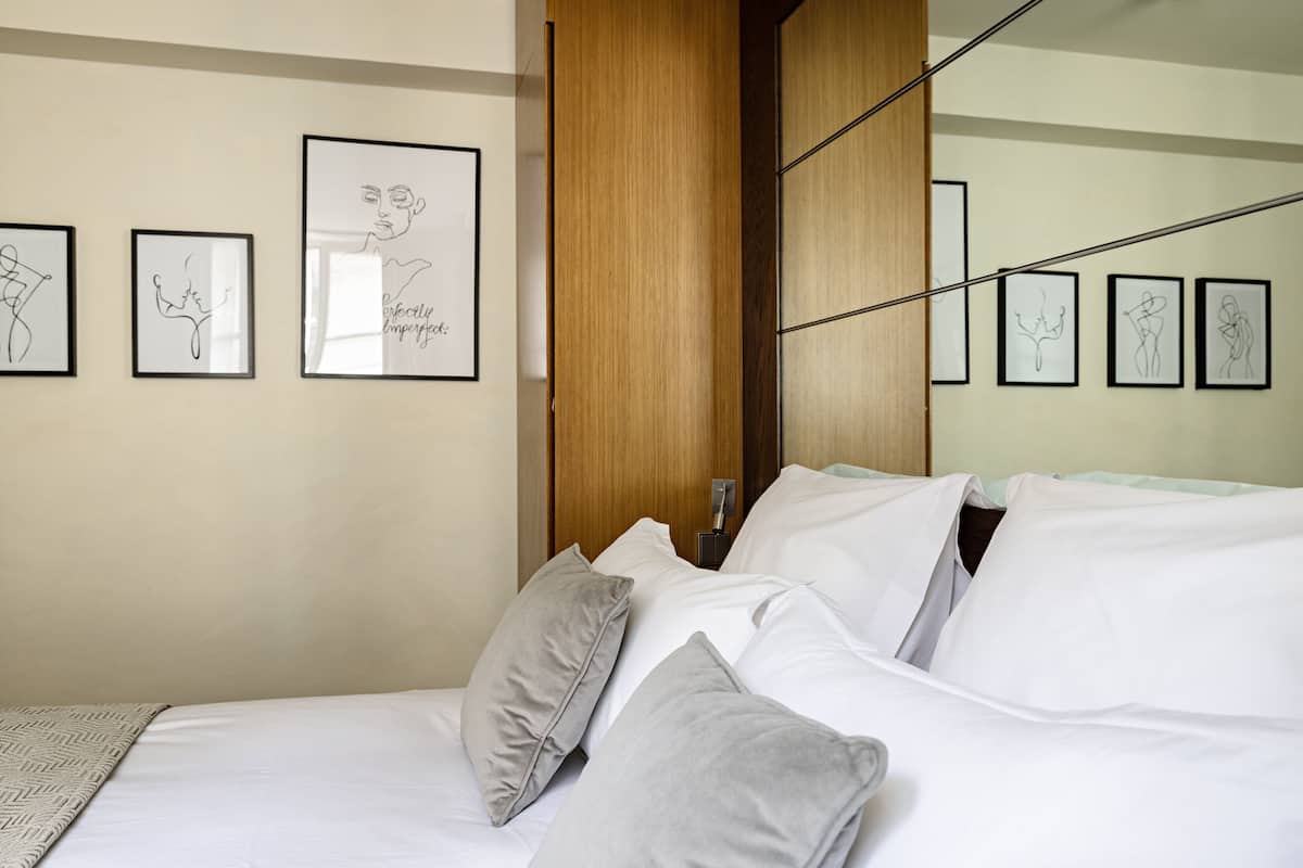 Un lit bien fait, avec du linge de lit blanc et des oreillers gris, un mur recouvert de panneaux en bois et des œuvres d’art encadrées accrochées au mur.