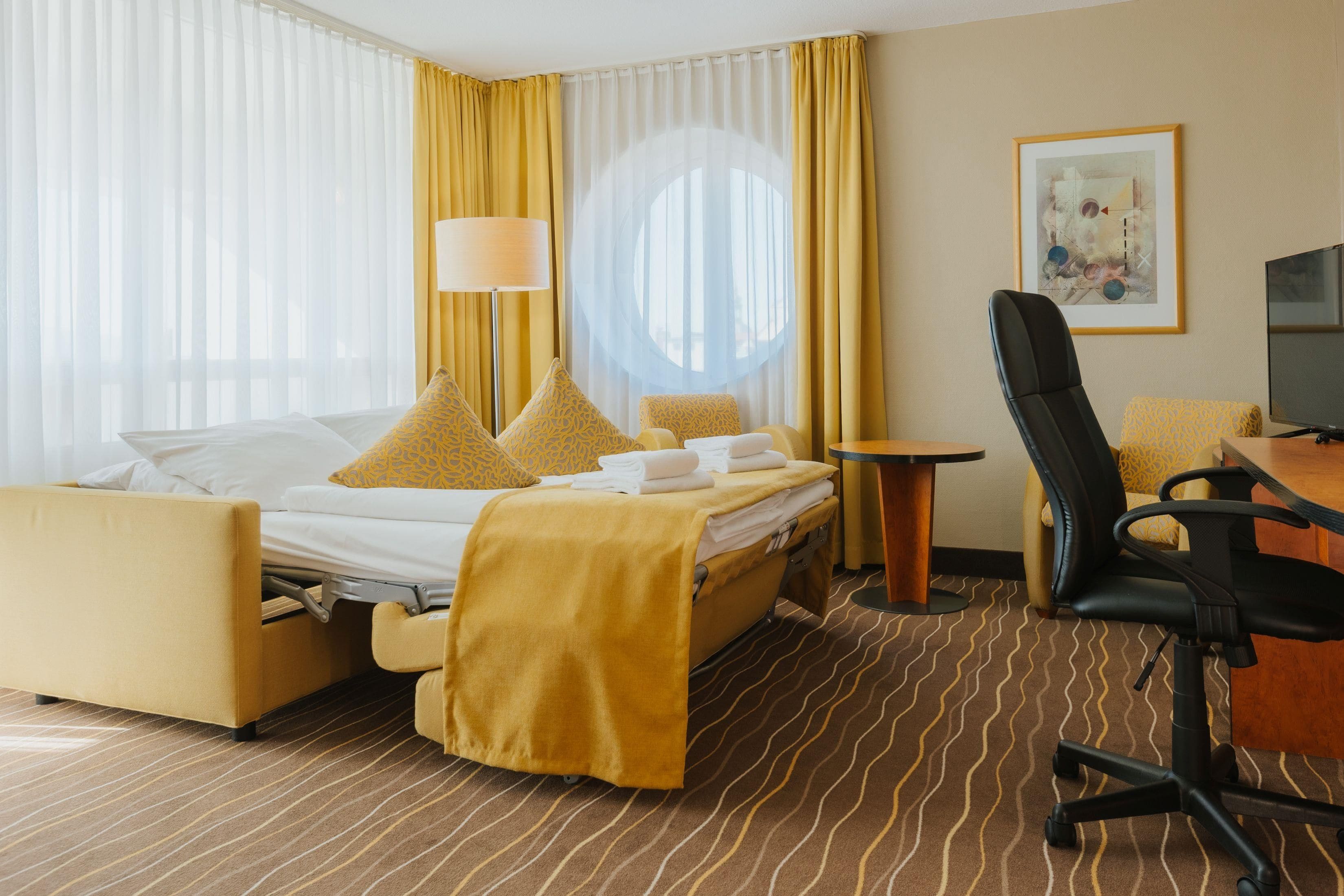 Ein Hotelzimmer mit einem Bett, einem Schreibtisch, einem Sessel, einem Fernseher und einem Vorhangfenster.