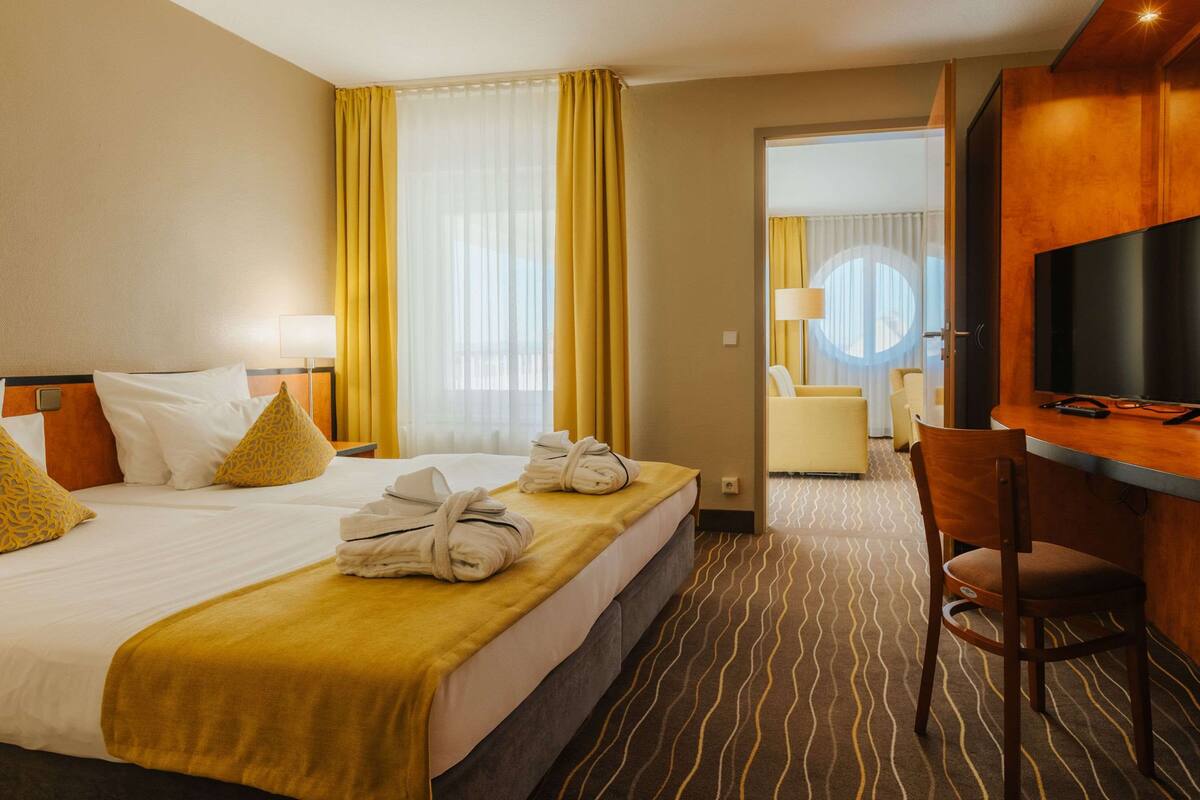 Quarto de hotel com cama, escrivaninha, poltrona, televisão e banheiro visível por uma porta aberta.