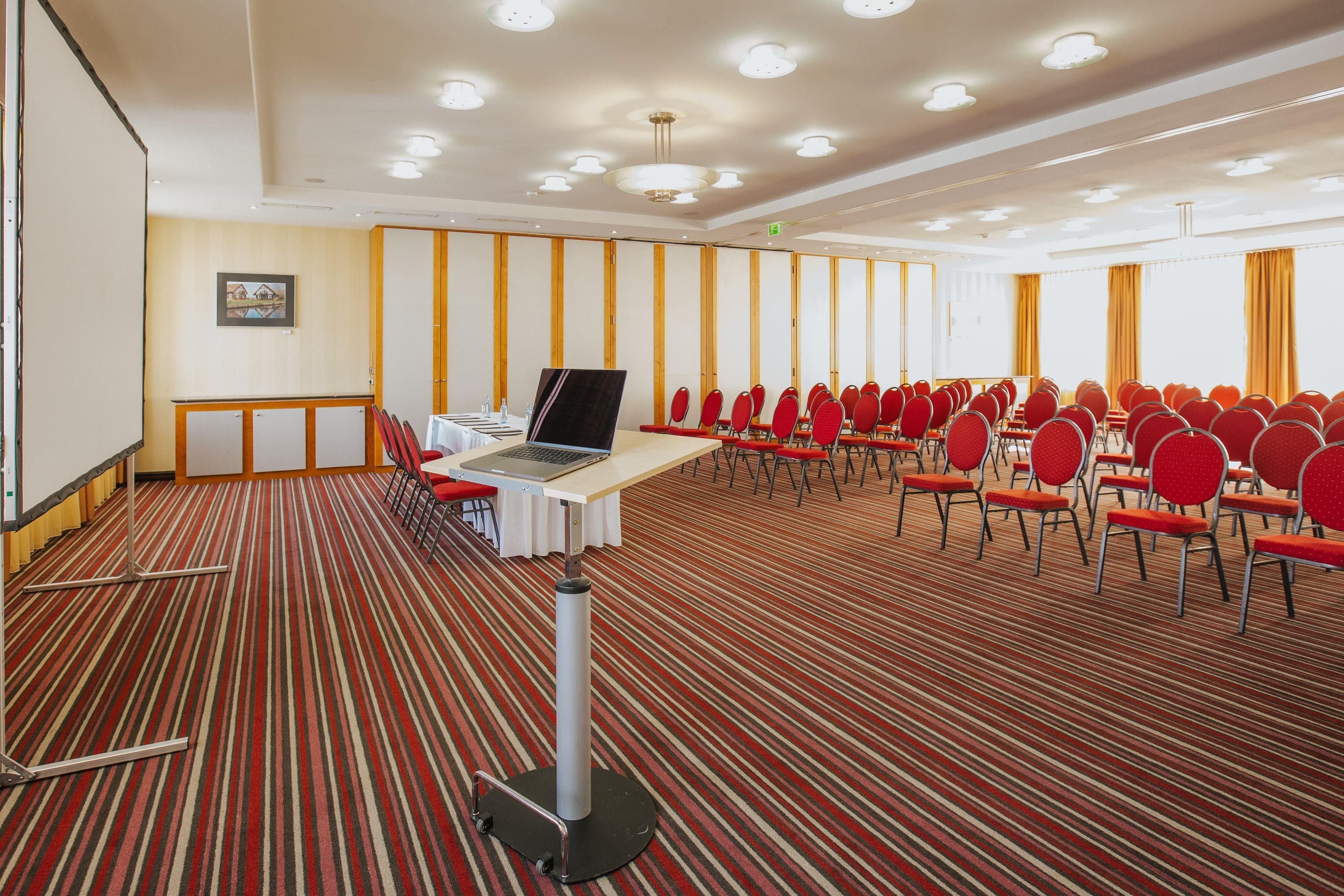 A conference room with a carpeted floor, a table with a laptop, and rows of red chairs.