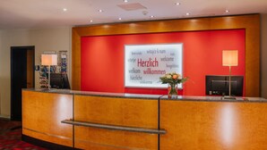A hotel reception desk with a red wall, a white welcome sign, and a vase with flowers. - Best Western Plus Hotel Bautzen (Bautzen)