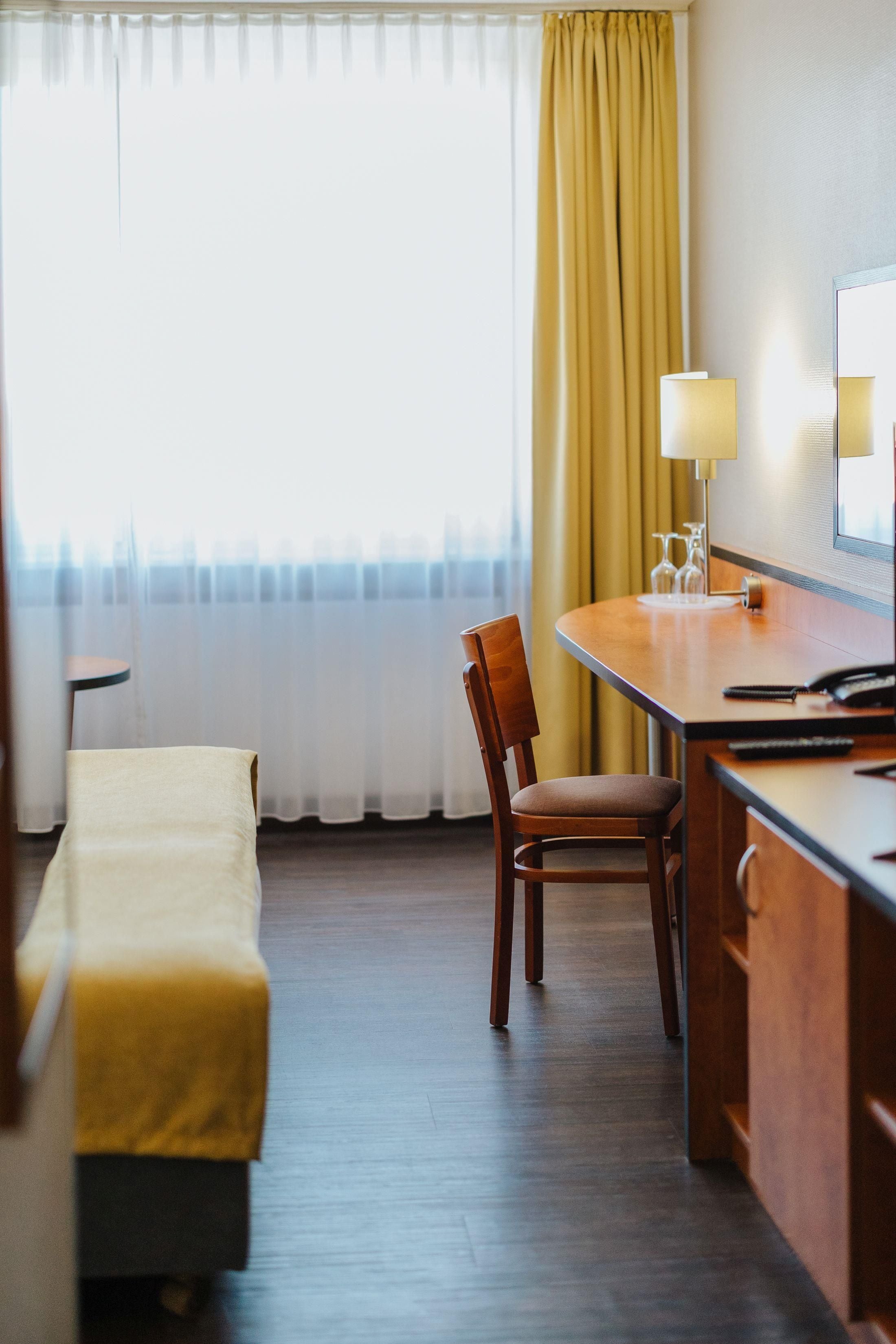 A hotel room with a wooden desk, a chair, a bed with a white cover, a window with curtains, and a lamp.