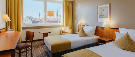 A hotel room with two beds, a desk, chairs, and a view of buildings through the window.