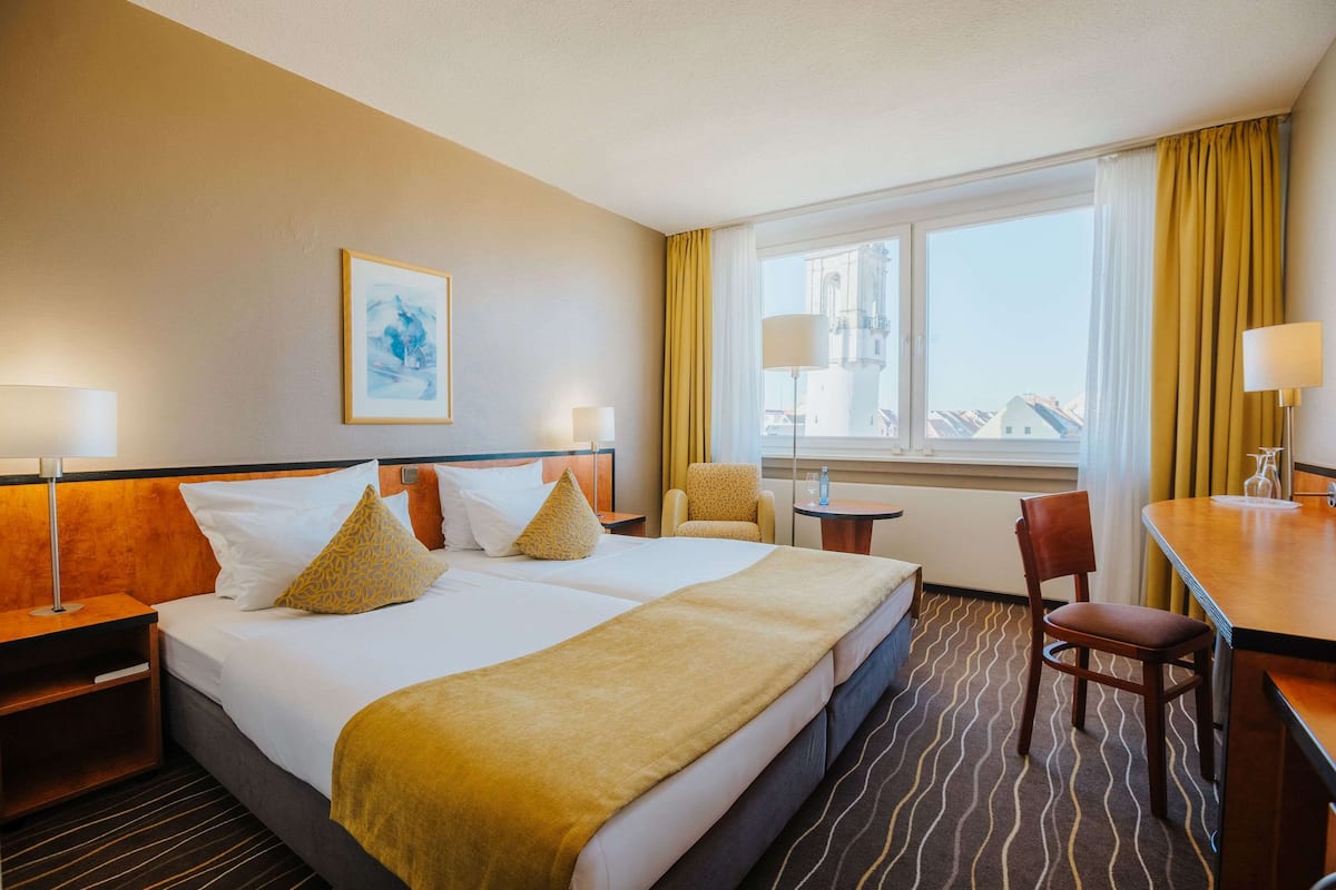 Quarto de hotel com uma cama grande, duas mesas de cabeceira, uma cadeira, uma escrivaninha e uma janela com cortinas.