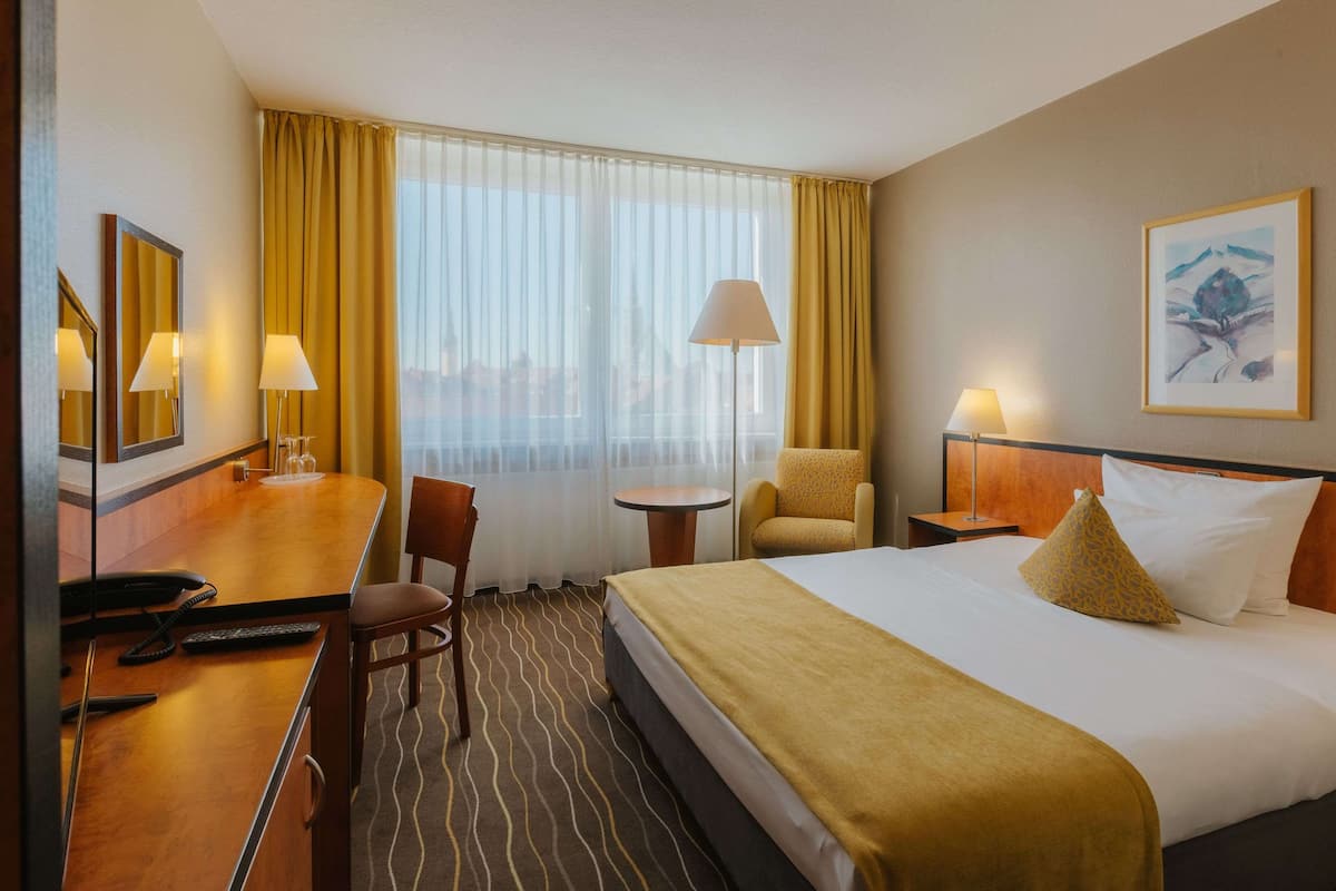 Quarto de hotel com cama, escrivaninha com telefone, cadeira, mesinha e abajur.