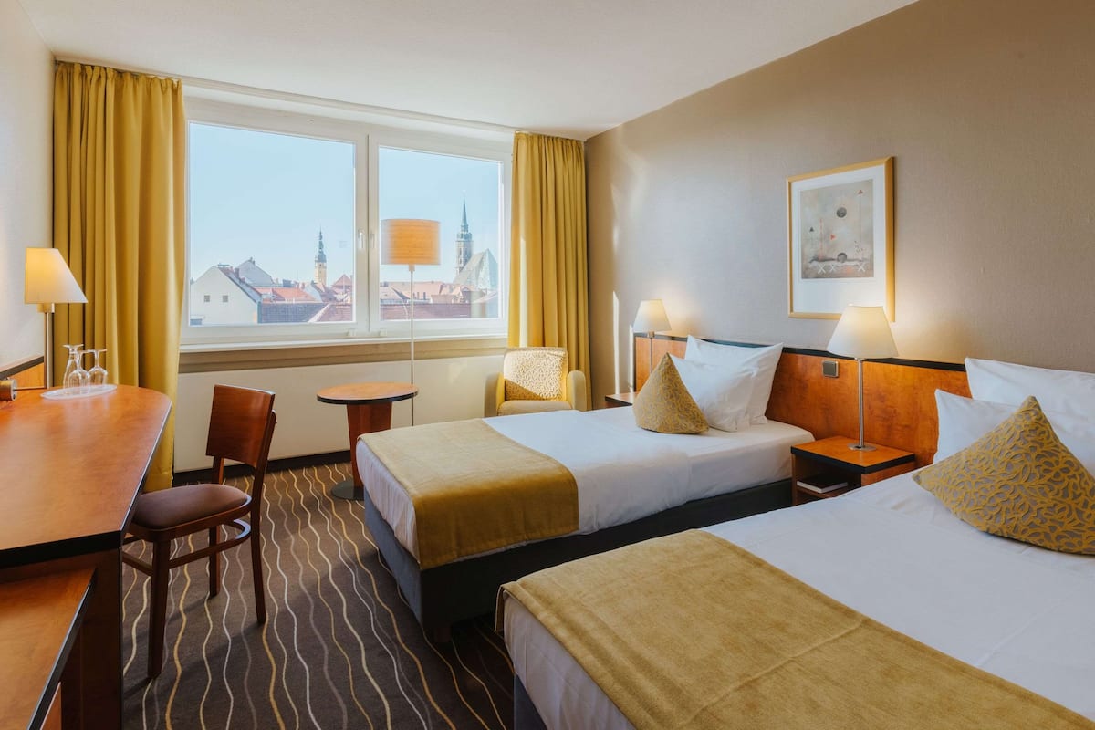 Quarto de hotel com duas camas, uma escrivaninha, uma cadeira, uma mesinha e uma janela com cortinas.
