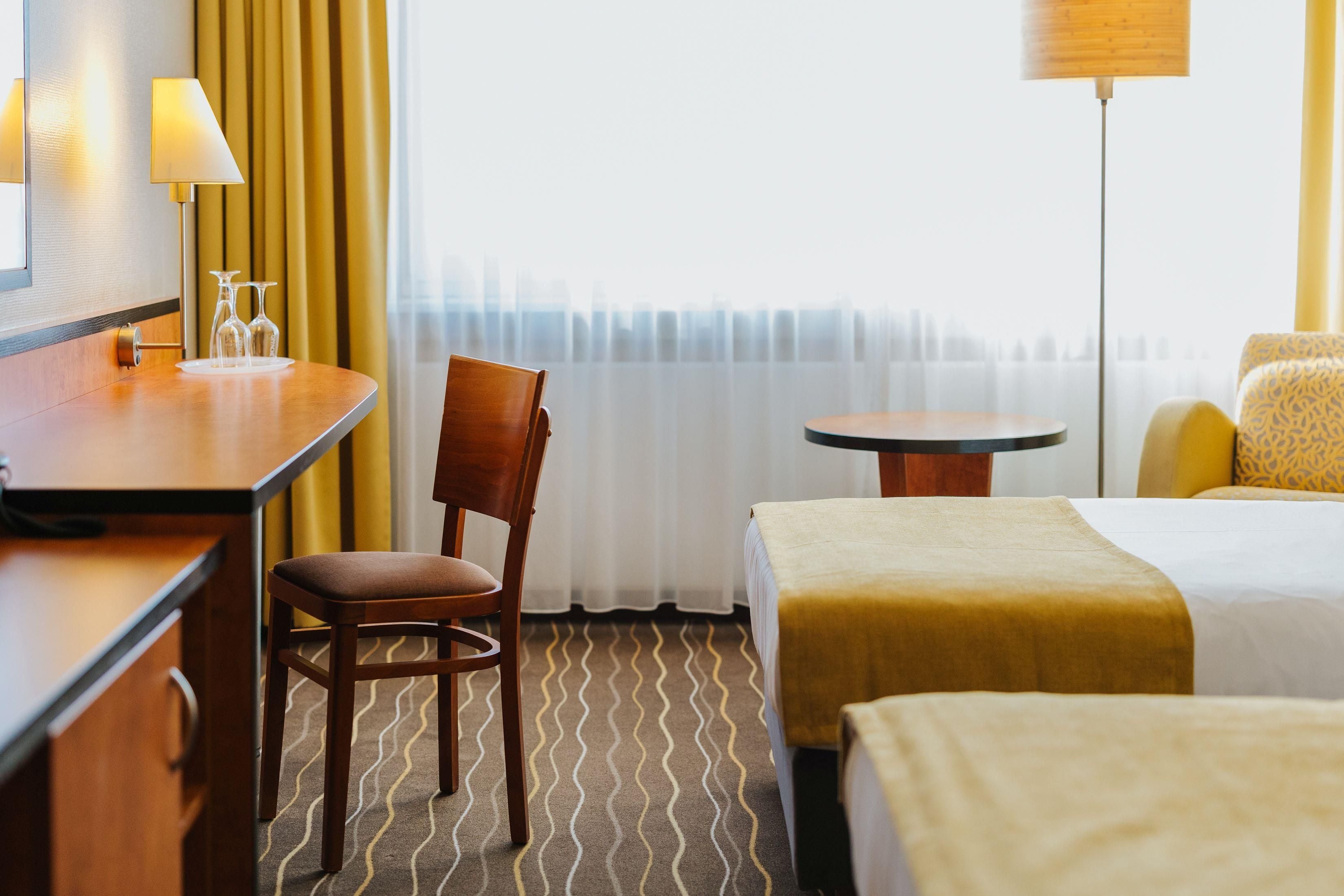 A hotel room with a bed, a desk, a chair, a small table, and a lamp.