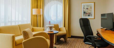 A hotel room with a sofa, armchairs, a small table with a bottle and glasses, a TV, and a painting on the wall.