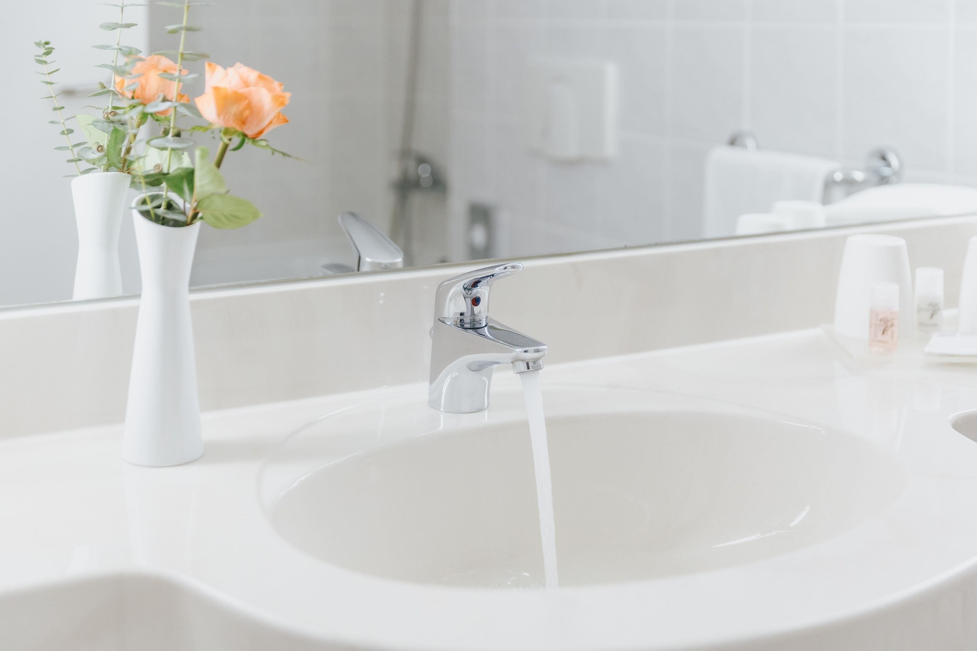 A bathroom sink with running water, a white vase with flowers, and a mirror reflecting the bathroom.