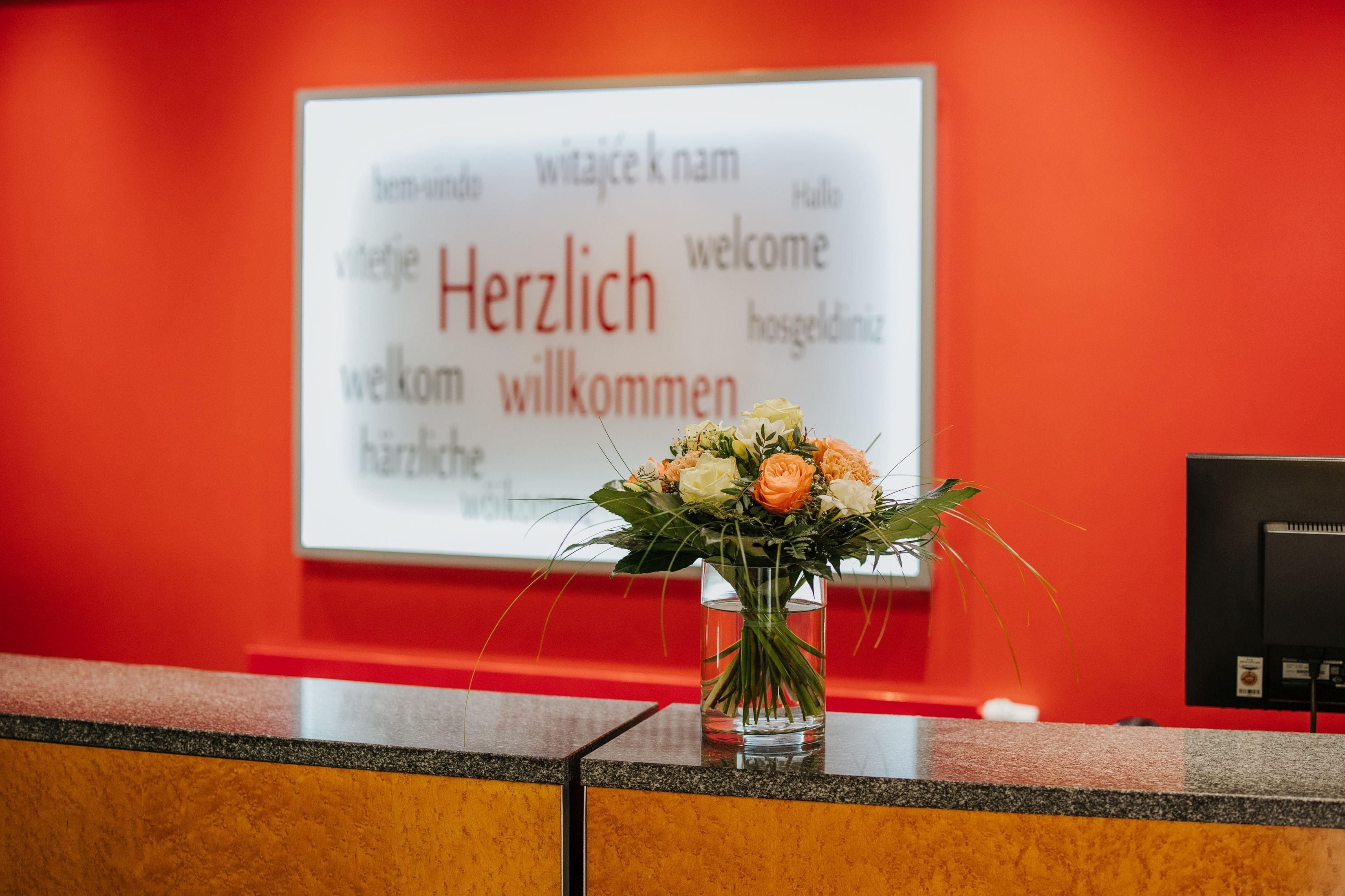 A reception desk with a welcome sign in multiple languages and a vase of flowers.