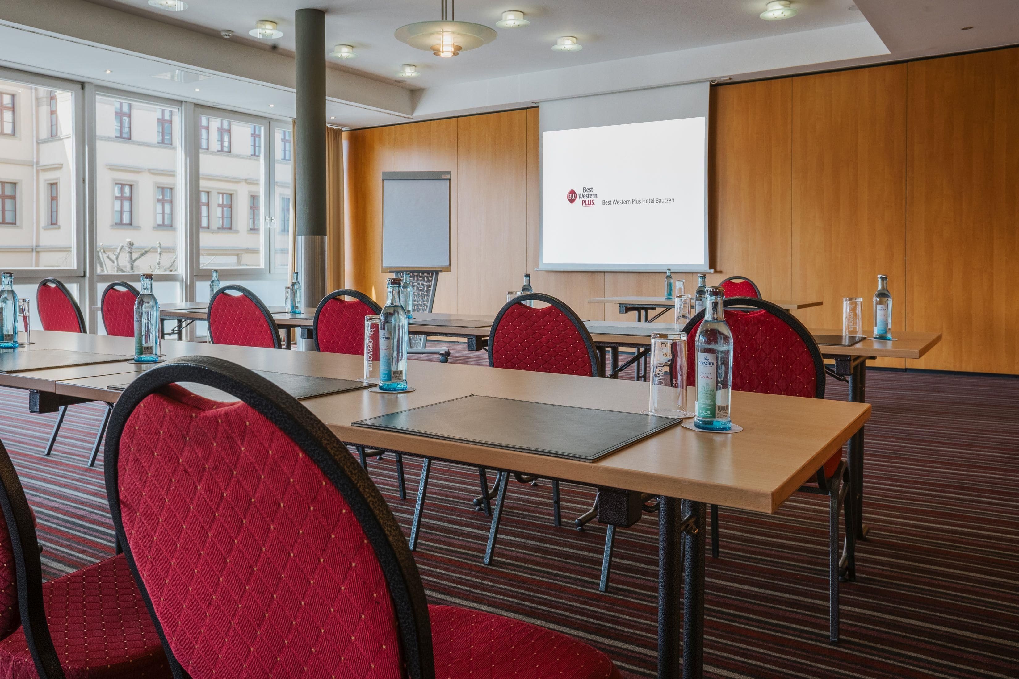 Ein Konferenzraum mit einer großen Leinwand, roten Polsterstühlen und Tischen mit Wasserflaschen.