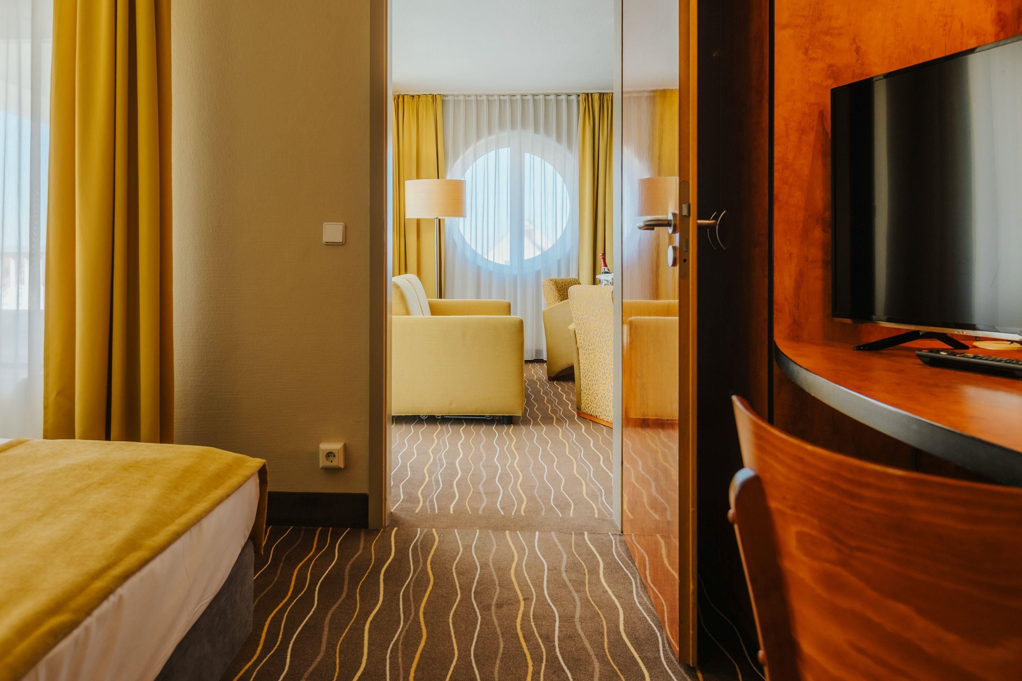 A hotel room with a bed, a desk with a TV, a seating area with two chairs, and a patterned carpet.