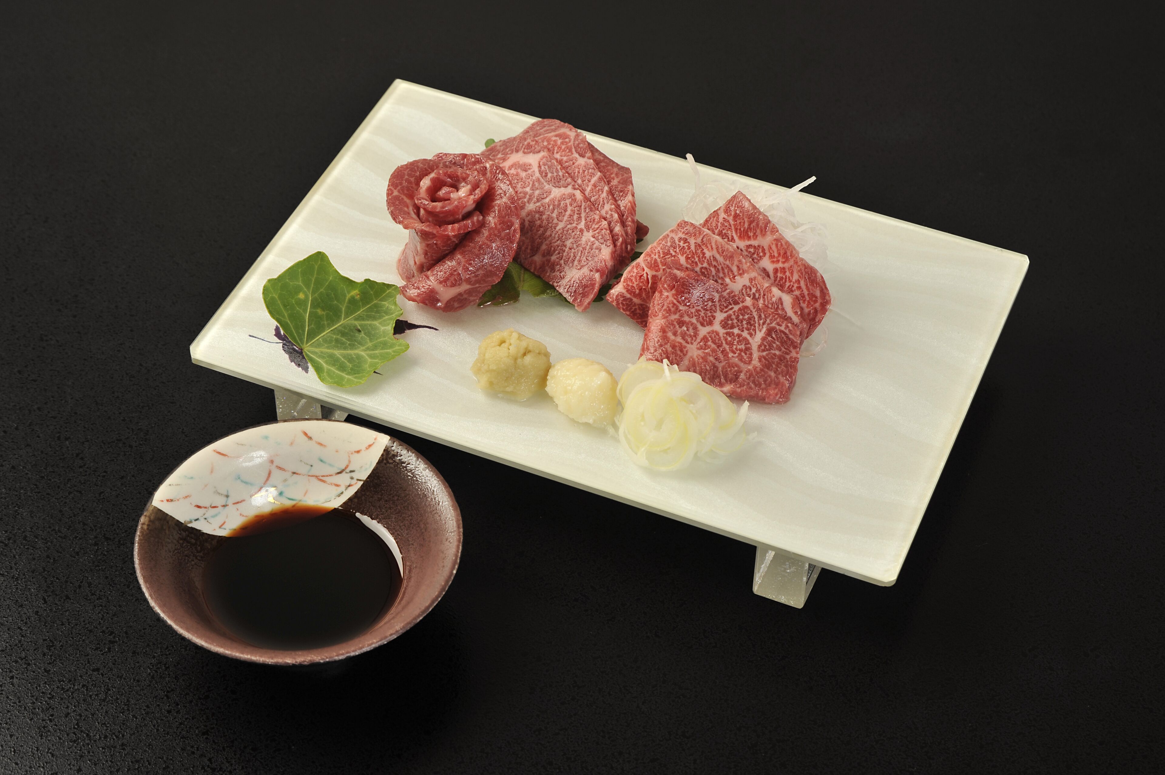 Une assiette rectangulaire blanche contenant différentes sortes de viande, une feuille et un petit bol de sauce.