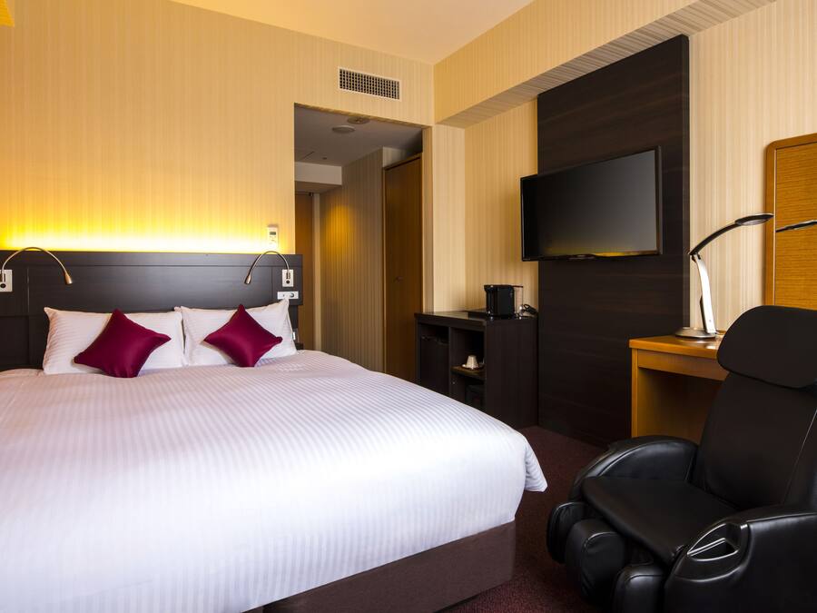 A hotel room with a large bed, a TV mounted on the wall, a massage chair, and a desk with a lamp.