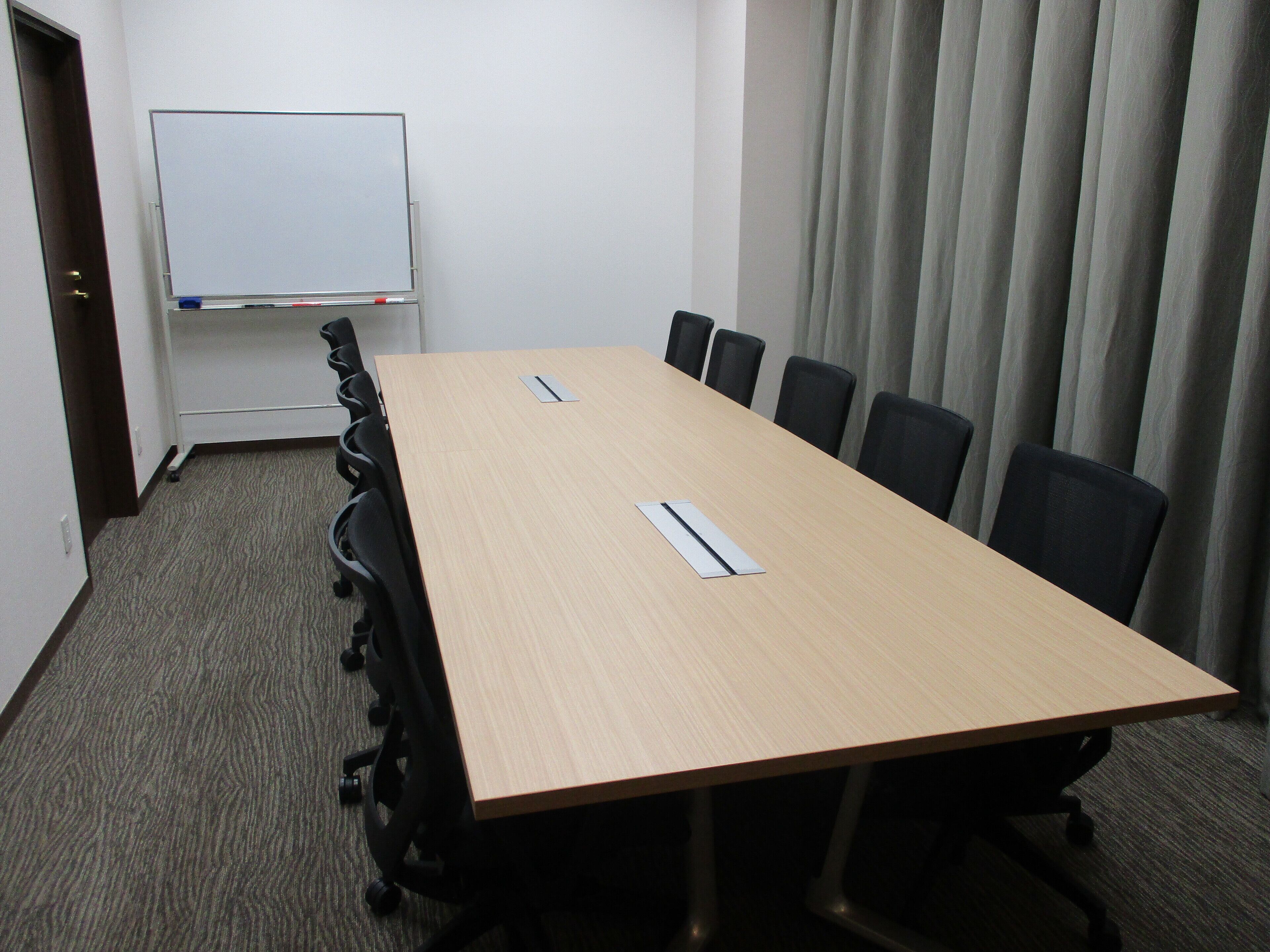 A conference room with a long table, six chairs, a whiteboard, and a curtain.