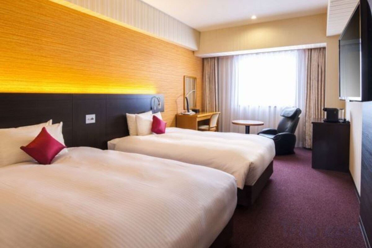 A hotel room with two beds, a desk, a chair, a TV, and a window with curtains.