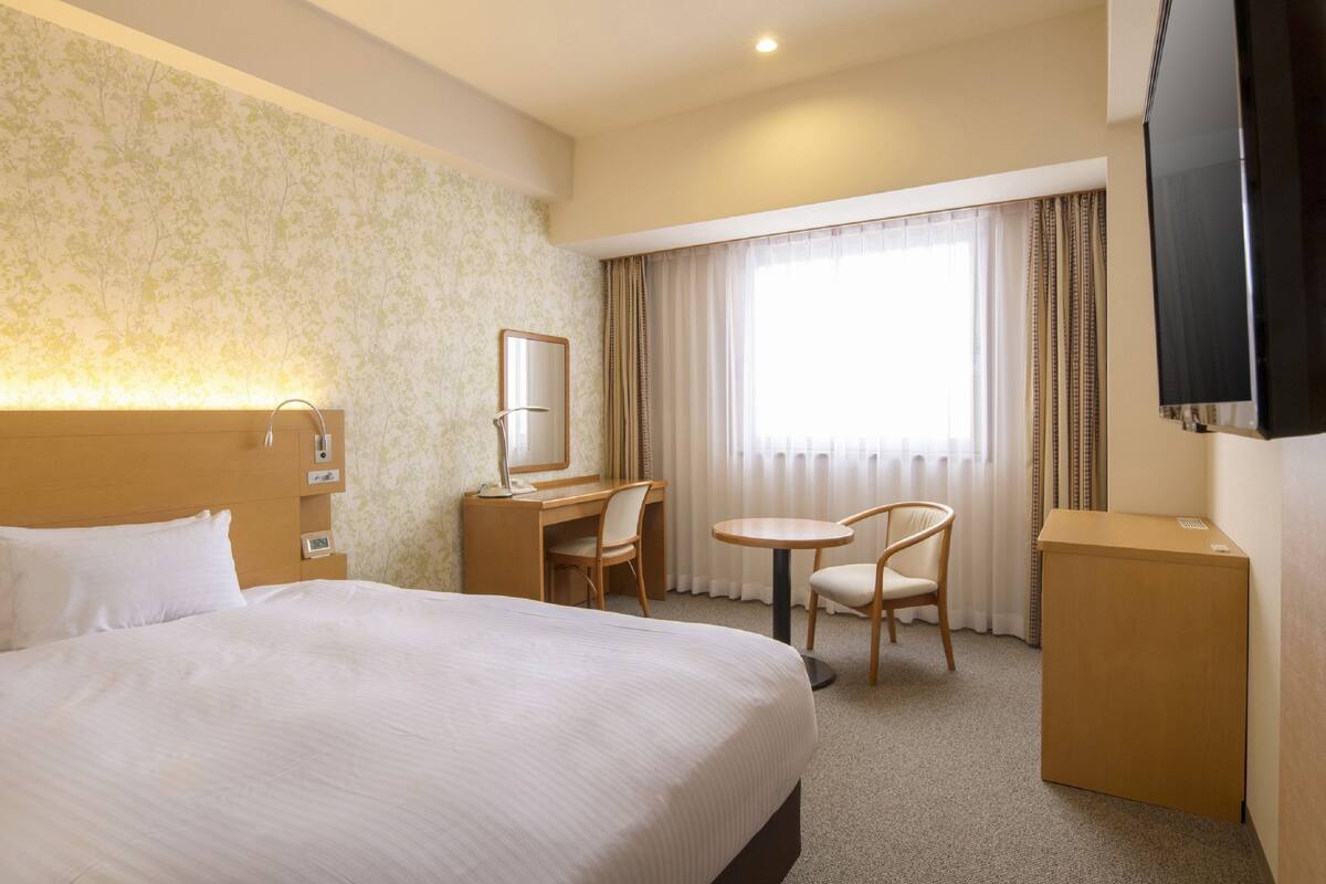 A hotel room with a large bed, a desk with a chair, a small table, a TV, and a window with curtains.