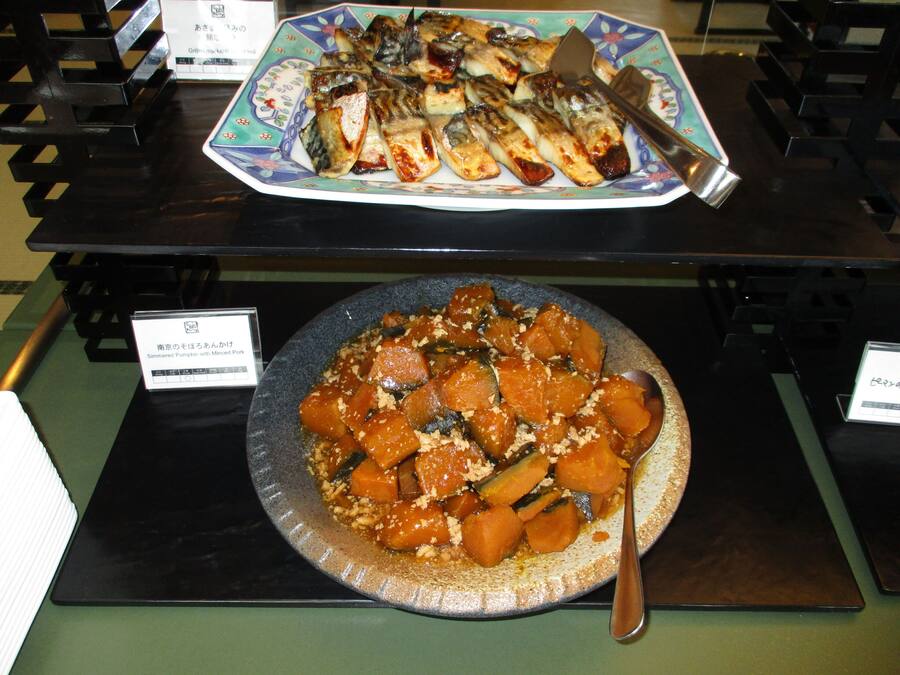 A buffet with a variety of dishes including a plate of grilled fish and a bowl of simmered pumpkin with minced pork.
