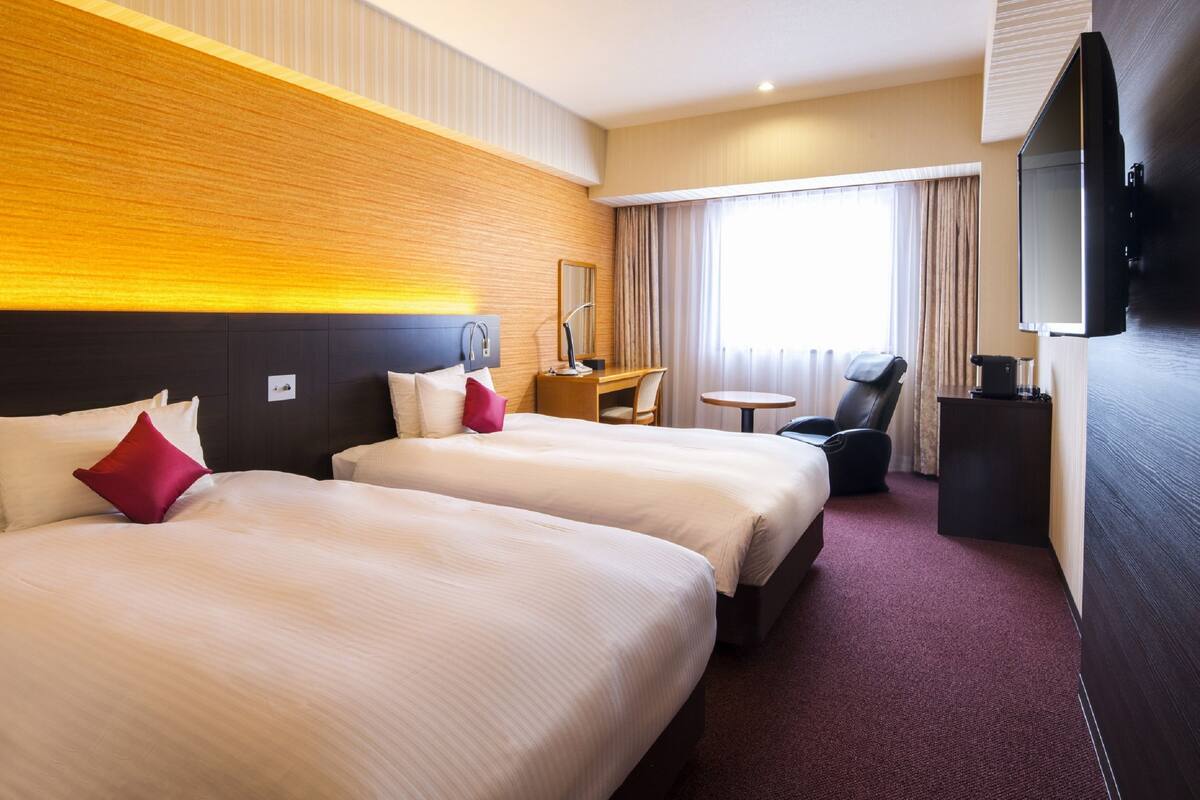 A hotel room with two beds, a desk, a chair, a TV, and a window with curtains.