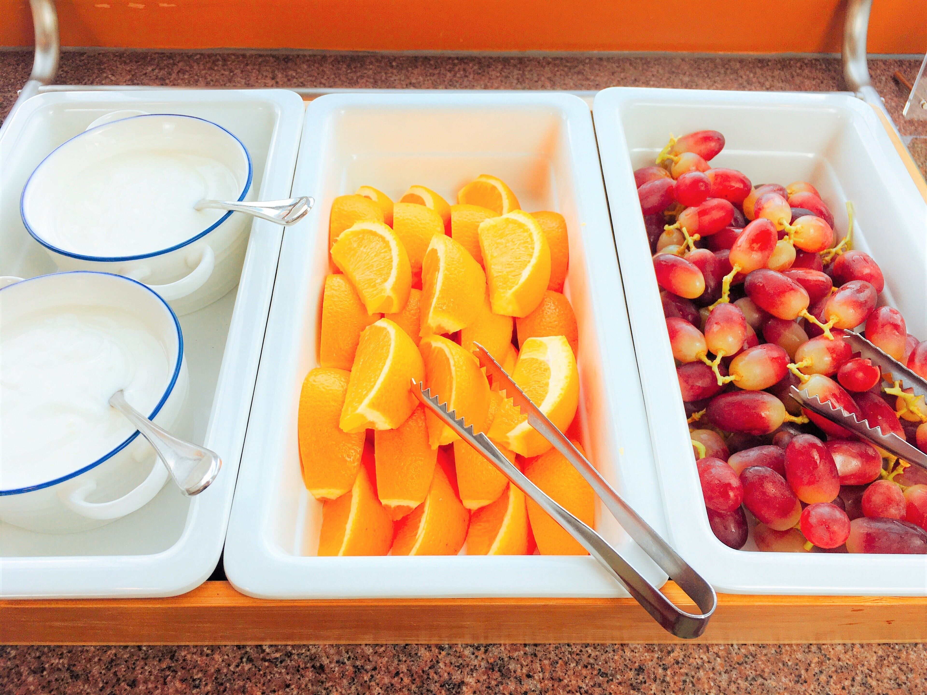 A buffet with three containers: one with orange slices, one with grapes, and one with a white creamy substance.