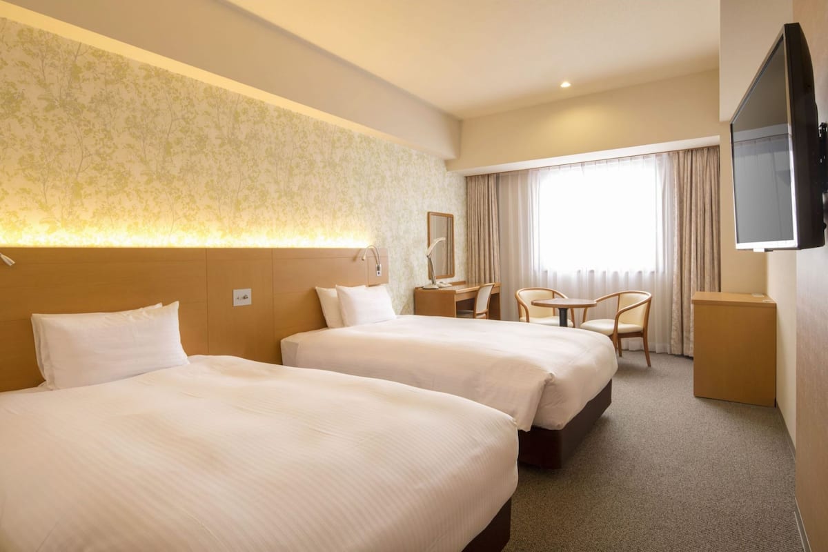 A hotel room with two beds, a desk, a chair, a TV, and a large window with curtains.